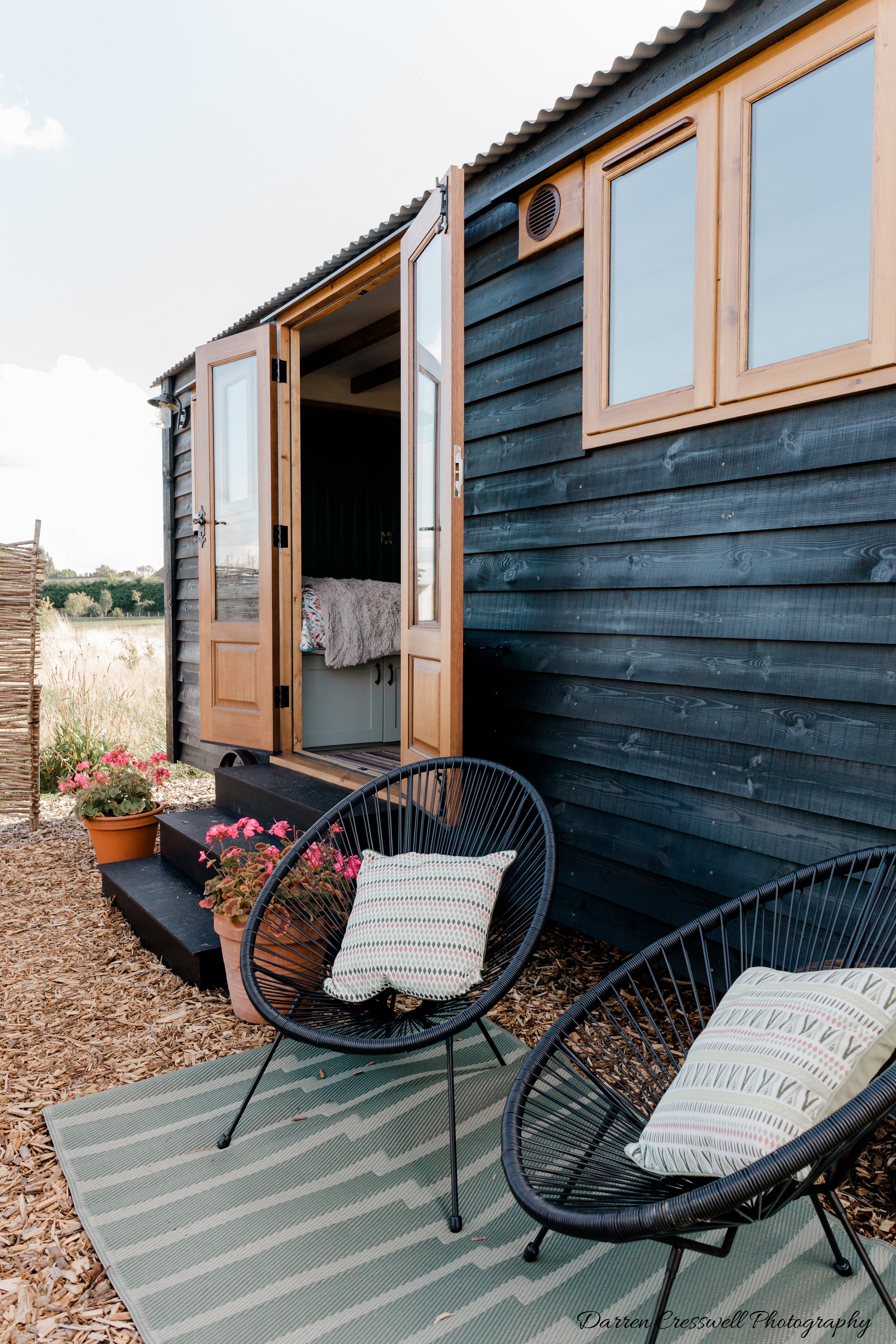 Outdoor patio scene with two black circular chairs with pillows, a green striped rug, potted flowers, and a small step leading to a black wooden tiny house with open door and window.