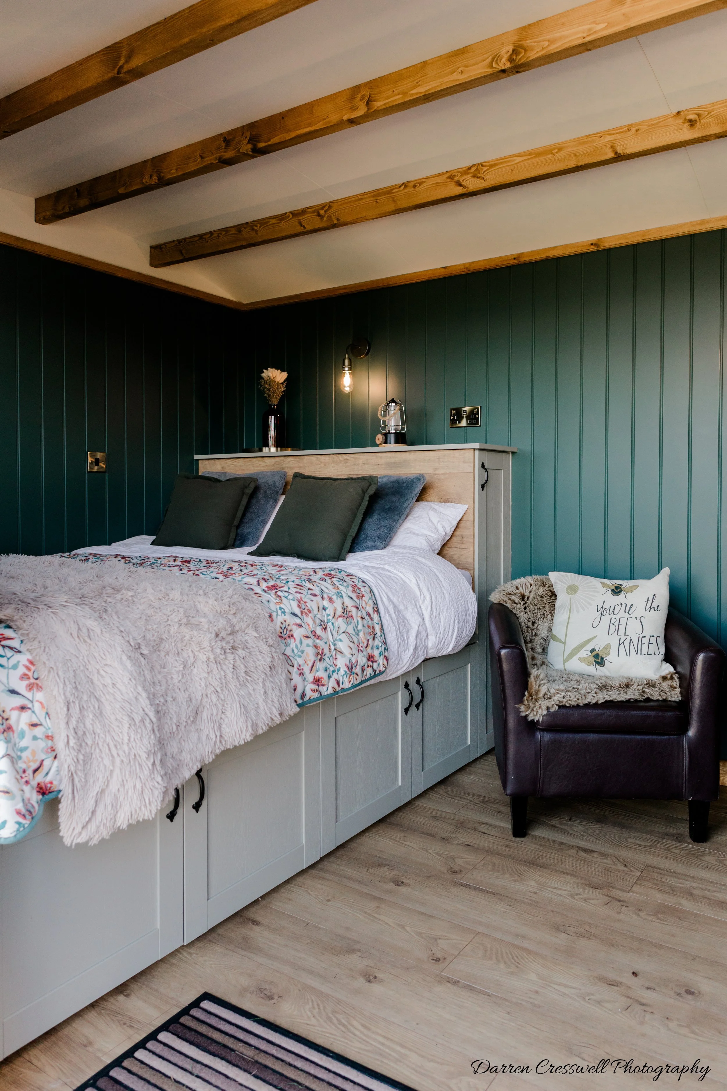 A bedroom with a bed featuring a floral bedspread, pillows, and a furry blanket, against a green paneled wall with wooden ceiling beams, with a black armchair with decorative pillows, a small side table, and wall-mounted lighting.