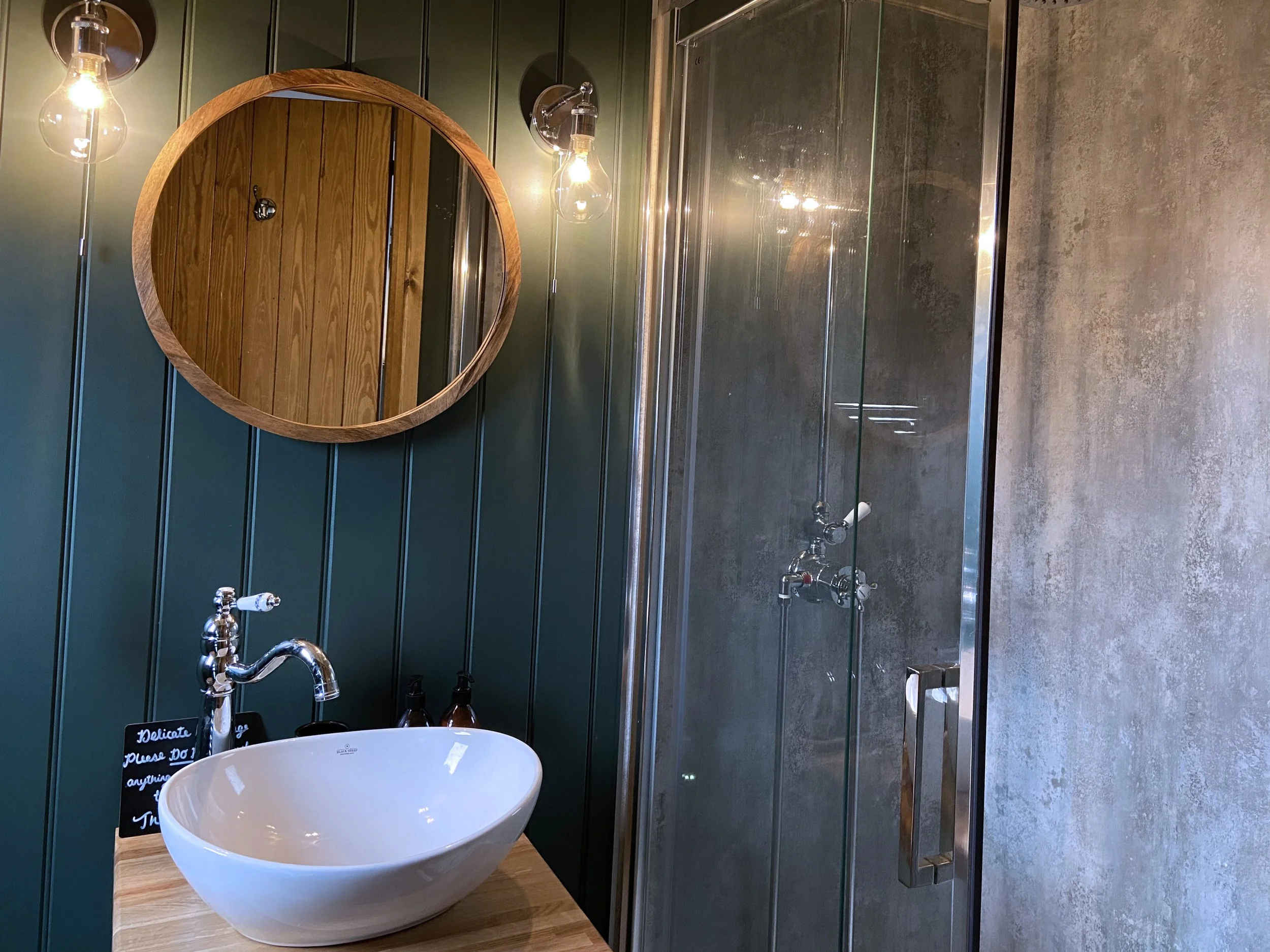A modern bathroom featuring a circular mirror above a white vessel sink with a vintage-style chrome faucet, illuminated by two exposed bulb wall sconces, with a shower enclosure on the right and green paneled wall behind the sink.