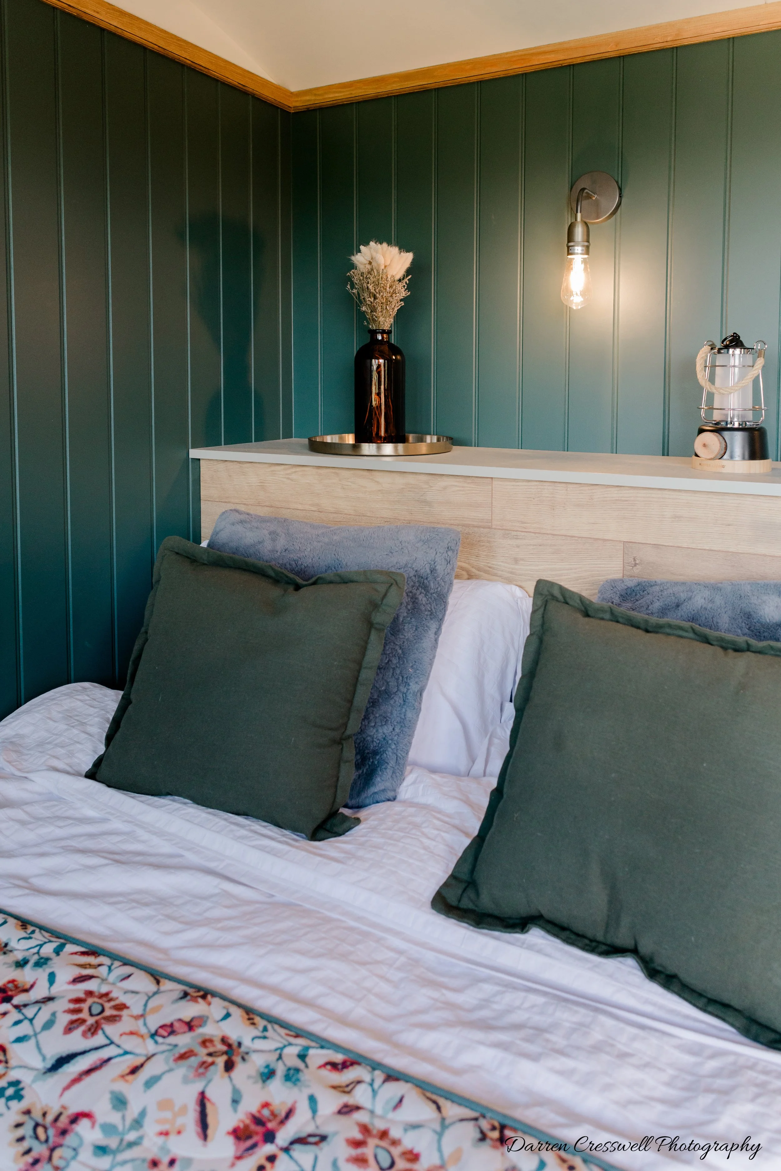 A bed with green and blue pillows against a wooden headboard, with a dark vase holding dried flowers, a wall sconce with an exposed lightbulb, and a small lamp on the nightstand in a room with green paneled walls.