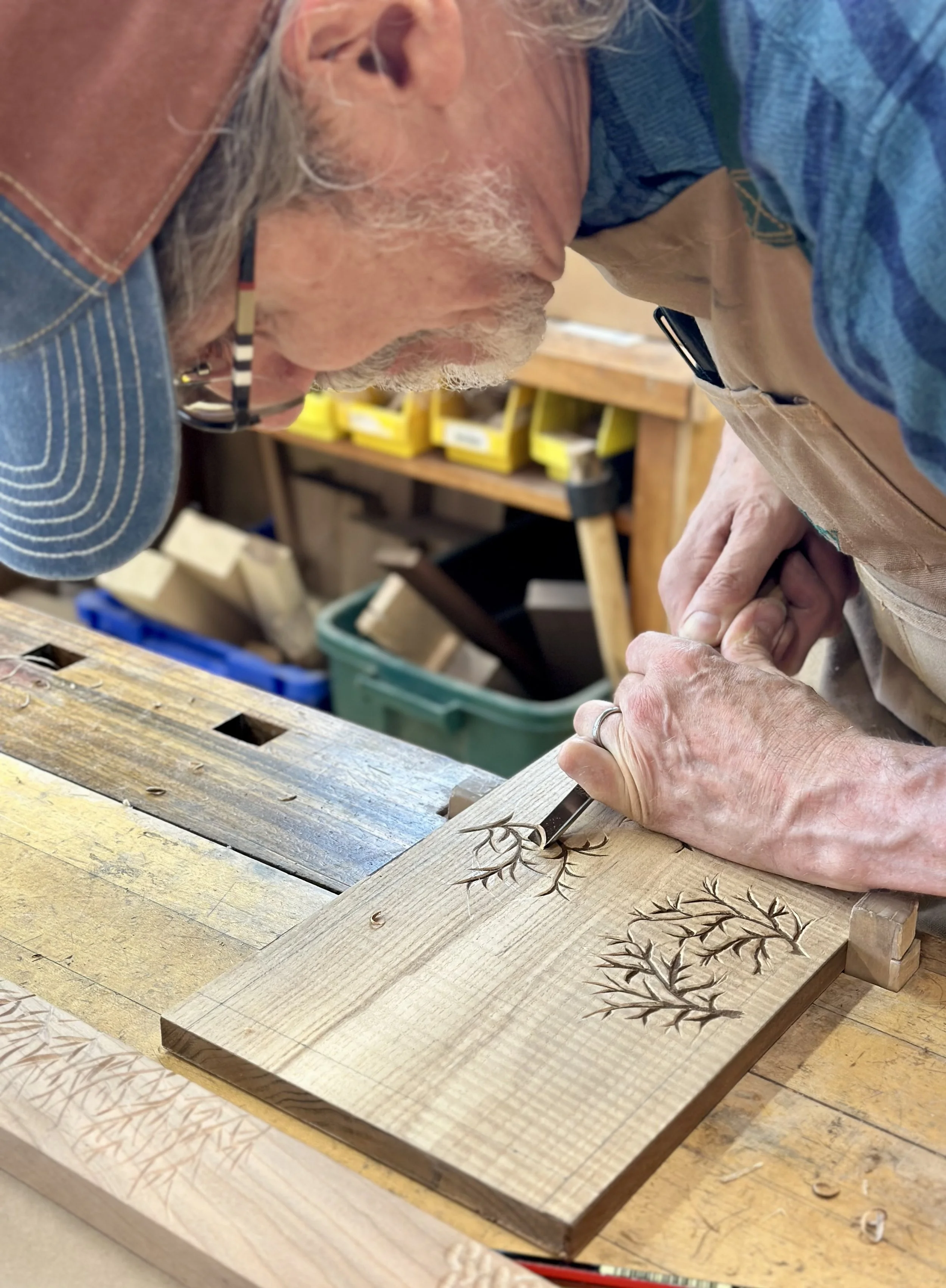 A person is carving a tree branch design into a piece of wood using a carving tool in a woodworking workshop.