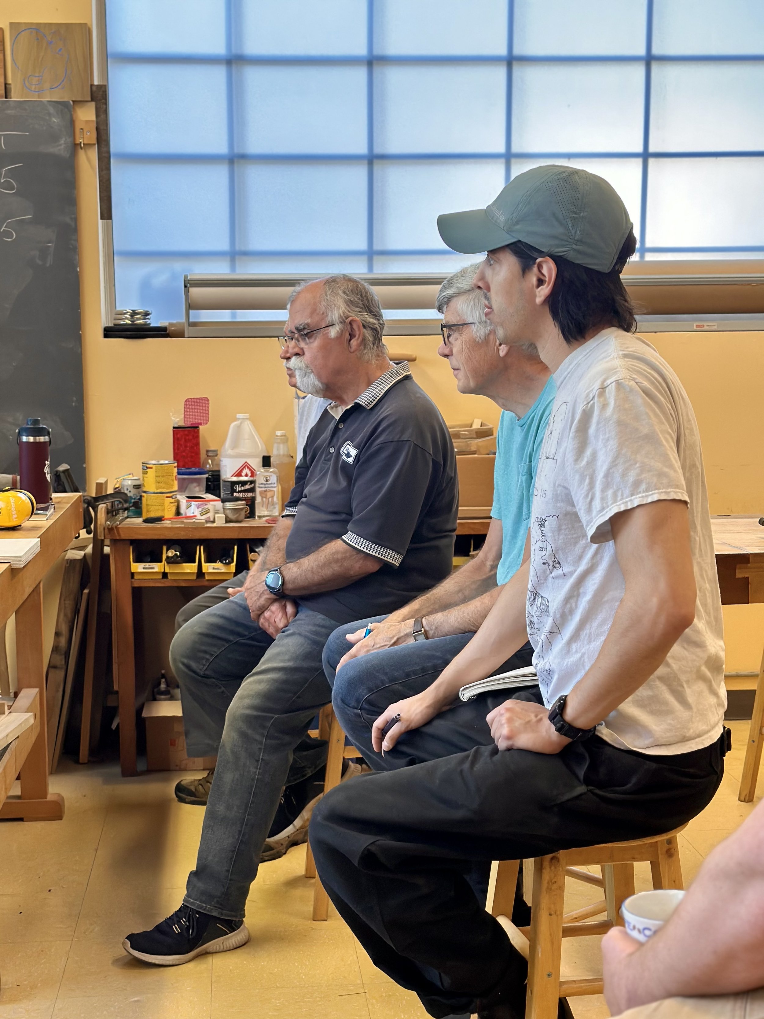 Four men sitting on stools in a workshop or classroom, facing to the right, with tools and supplies on a workbench behind them and a large window in the background.