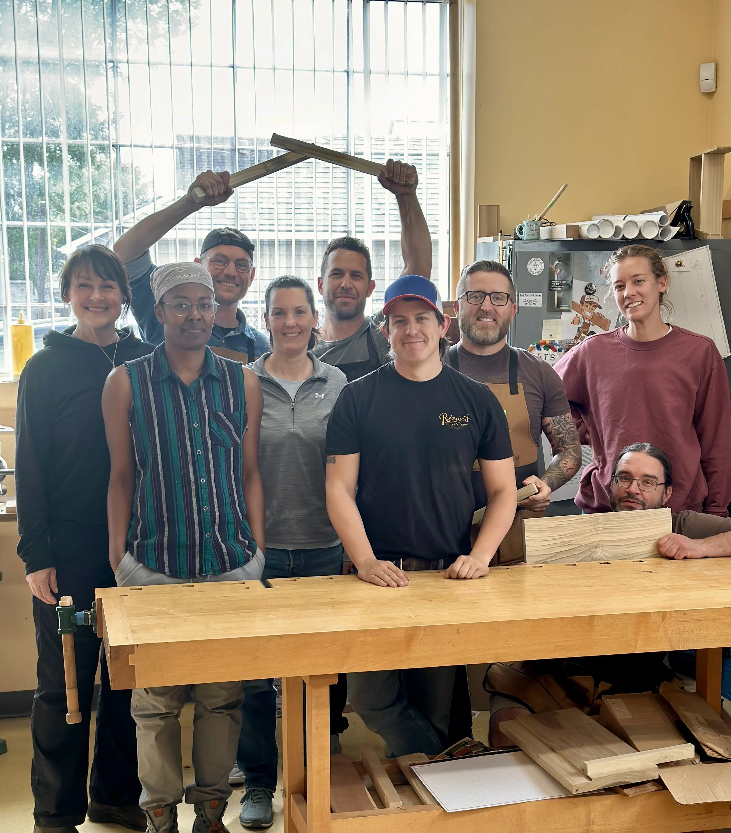 Group of nine people in a woodworking shop with some woodworking tools and materials, smiling at the camera. One person is holding a woodworking frame above his head.