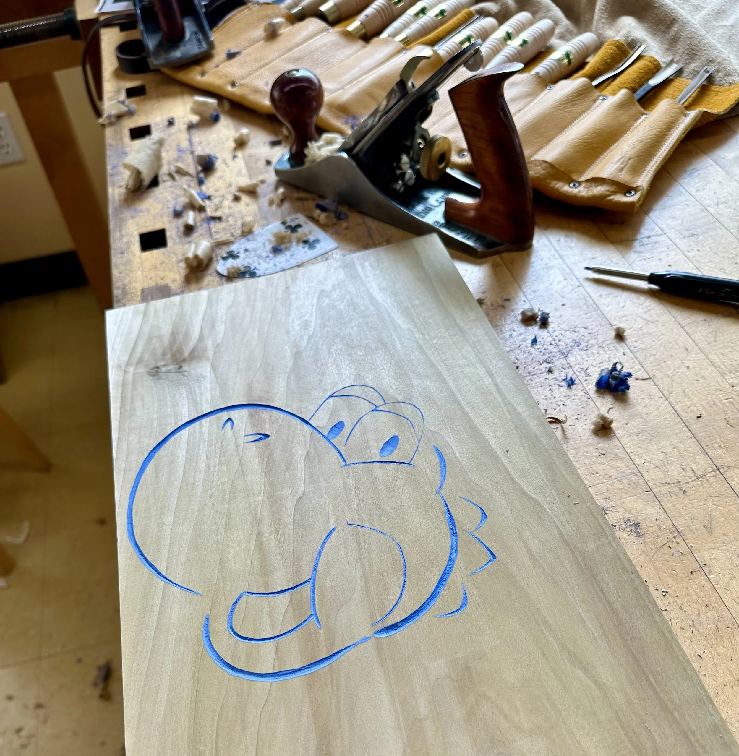 A woodworking workbench with a hand plane, a stack of rolled yellow leather pieces, a screw driver, a pen, and shavings and blue string on the surface.