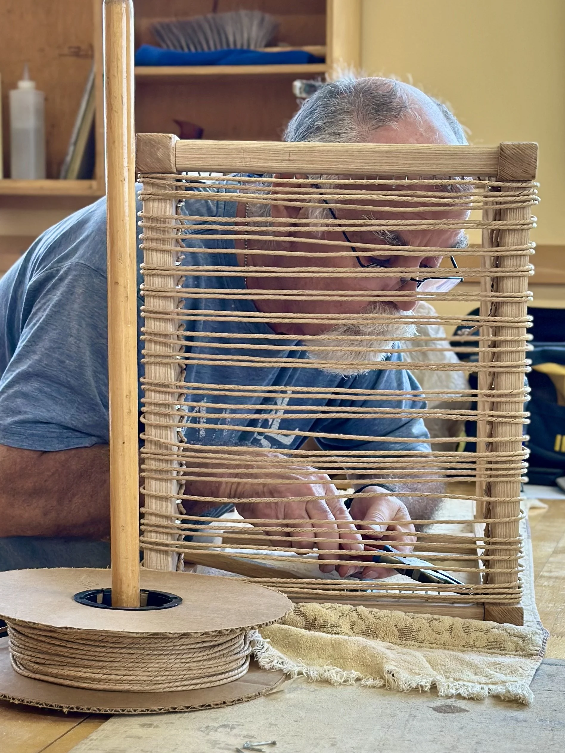 An elderly man with glasses working on a wicker furniture piece, behind a large woven wicker frame, with a wooden shelf in the background.