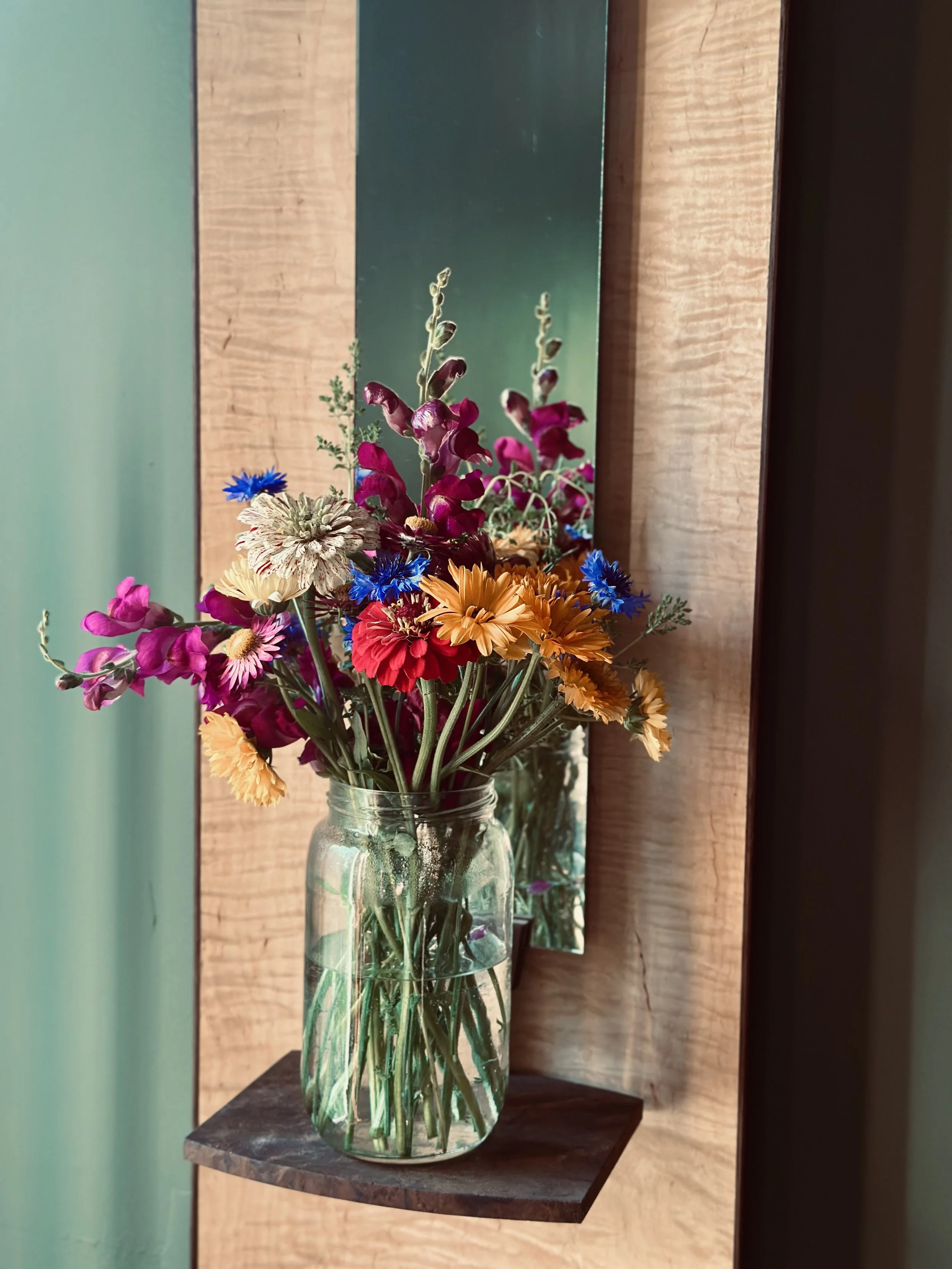 A glass vase with a colorful bouquet of flowers placed on a wooden surface against a wooden wall and a mint green wall.