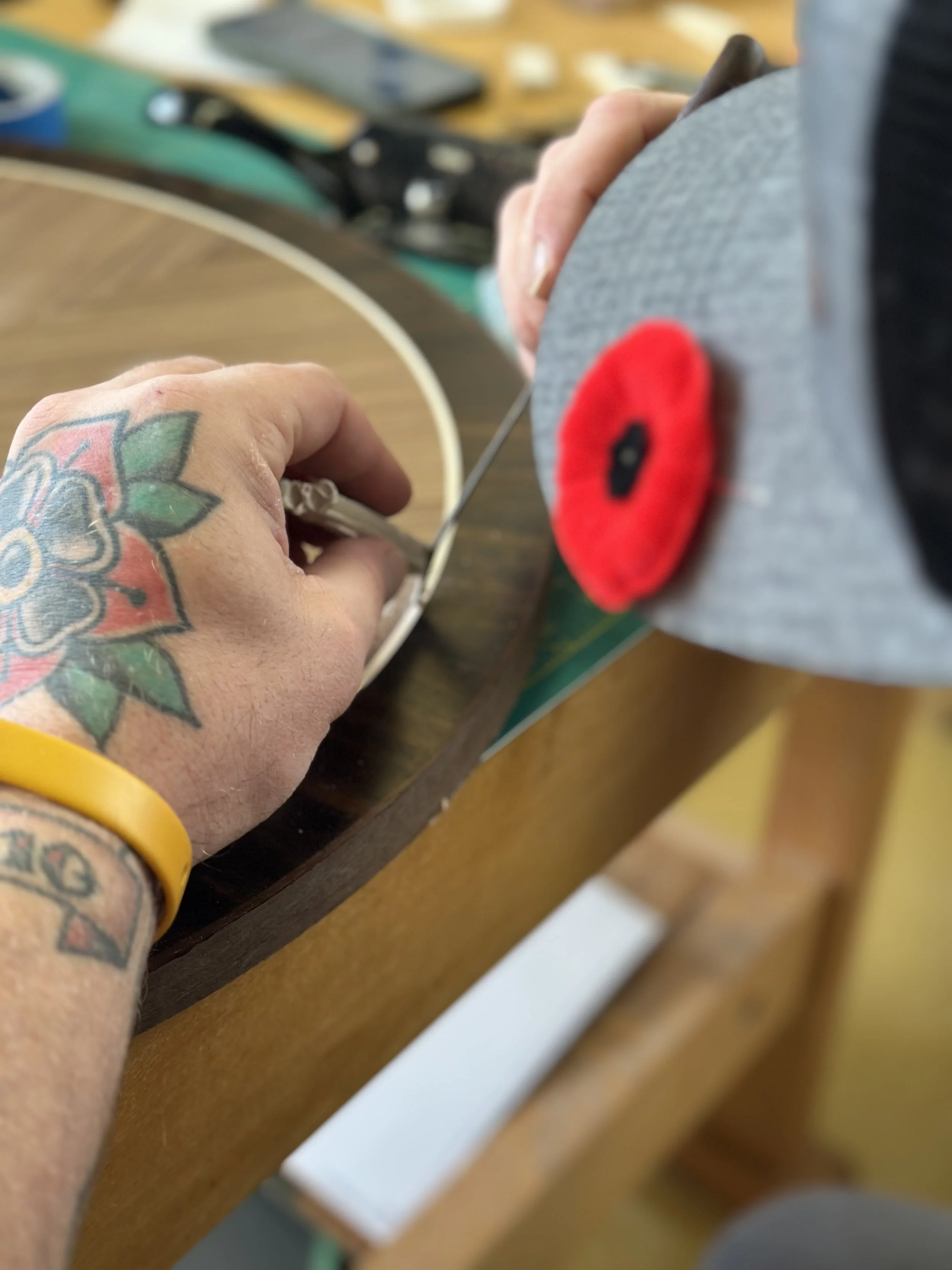 A person's hand with colorful tattoos holds a gray object with a red poppy pin. The background shows a wooden surface, a smartphone, and some tools.