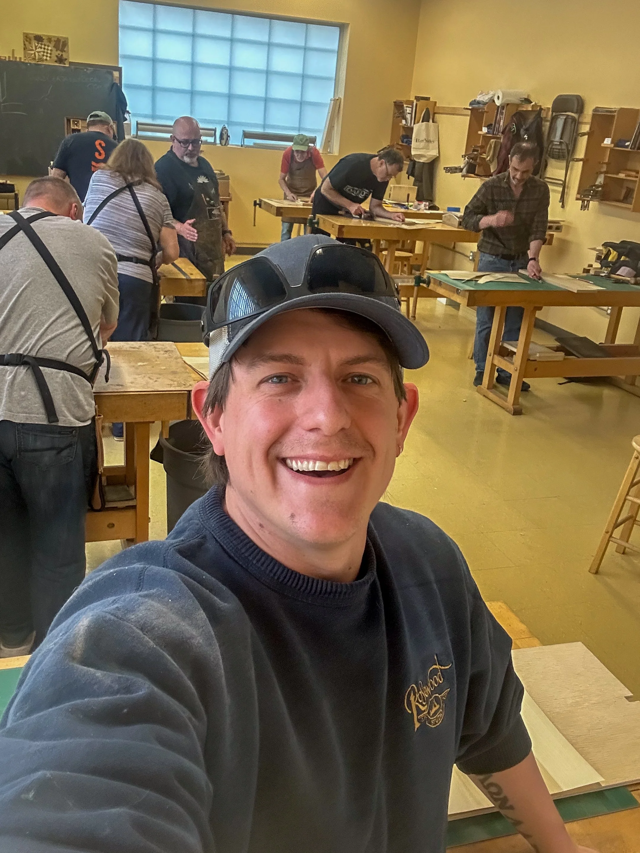 A group of people working on woodworking projects in a classroom or workshop, with tools and supplies on tables and shelves, and a man taking a selfie in the foreground.