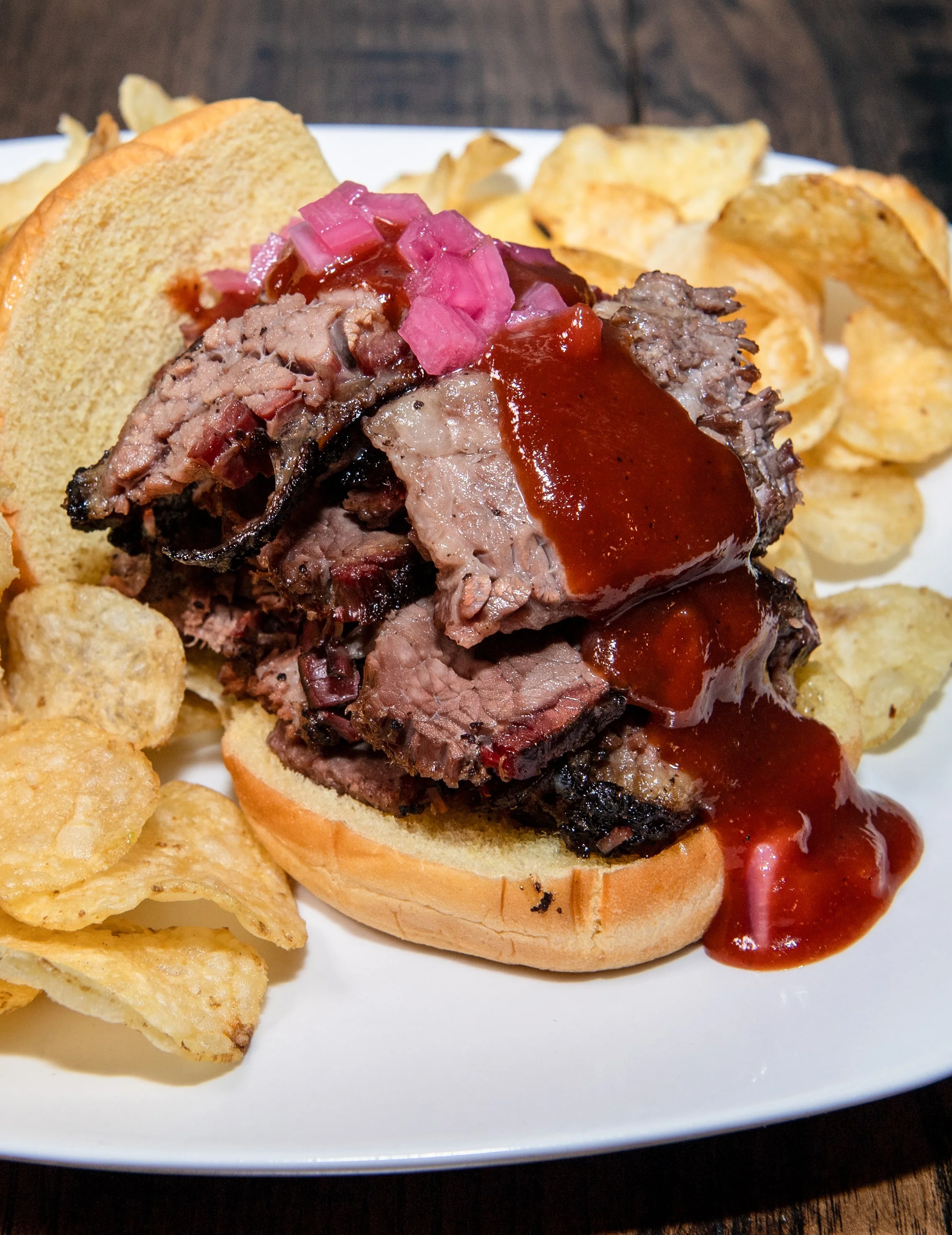 Barbecue beef brisket sandwich with pickled onions on a bun, served with potato chips on a white plate.