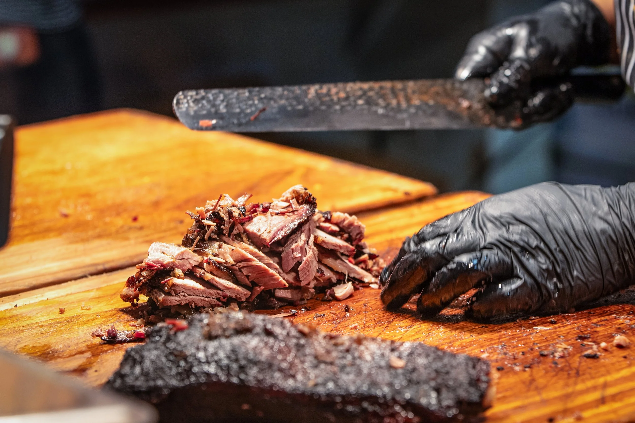 A person wearing black gloves is slicing smoked brisket on a wooden cutting board.