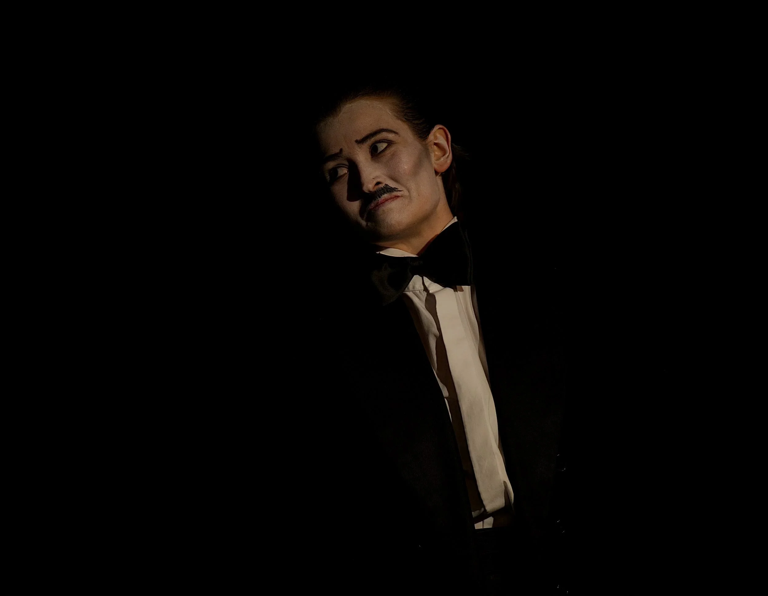 Person dressed as Charlie Chaplin with mustache, bow tie, and formal jacket, in a dark setting.