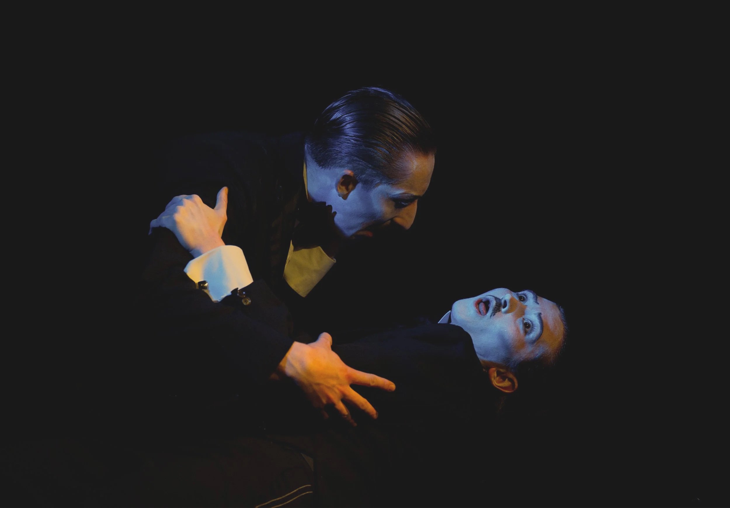 Two performers in black costumes with white face makeup, dark hair slicked back, are engaged in a theatrical scene against a black background. One performer leans over the other, who appears surprised or shocked.
