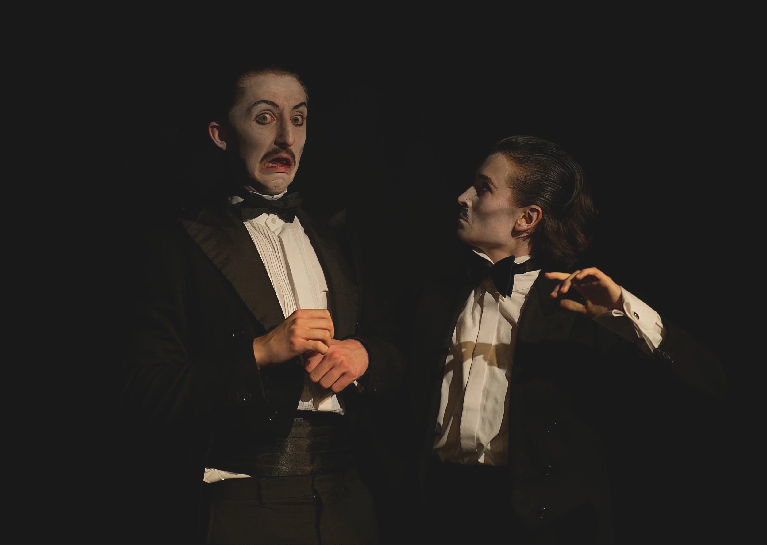 Two performers in tuxedos with bow ties, black coats, and white shirts, acting with exaggerated facial expressions against a black background, possibly during a theatrical performance.