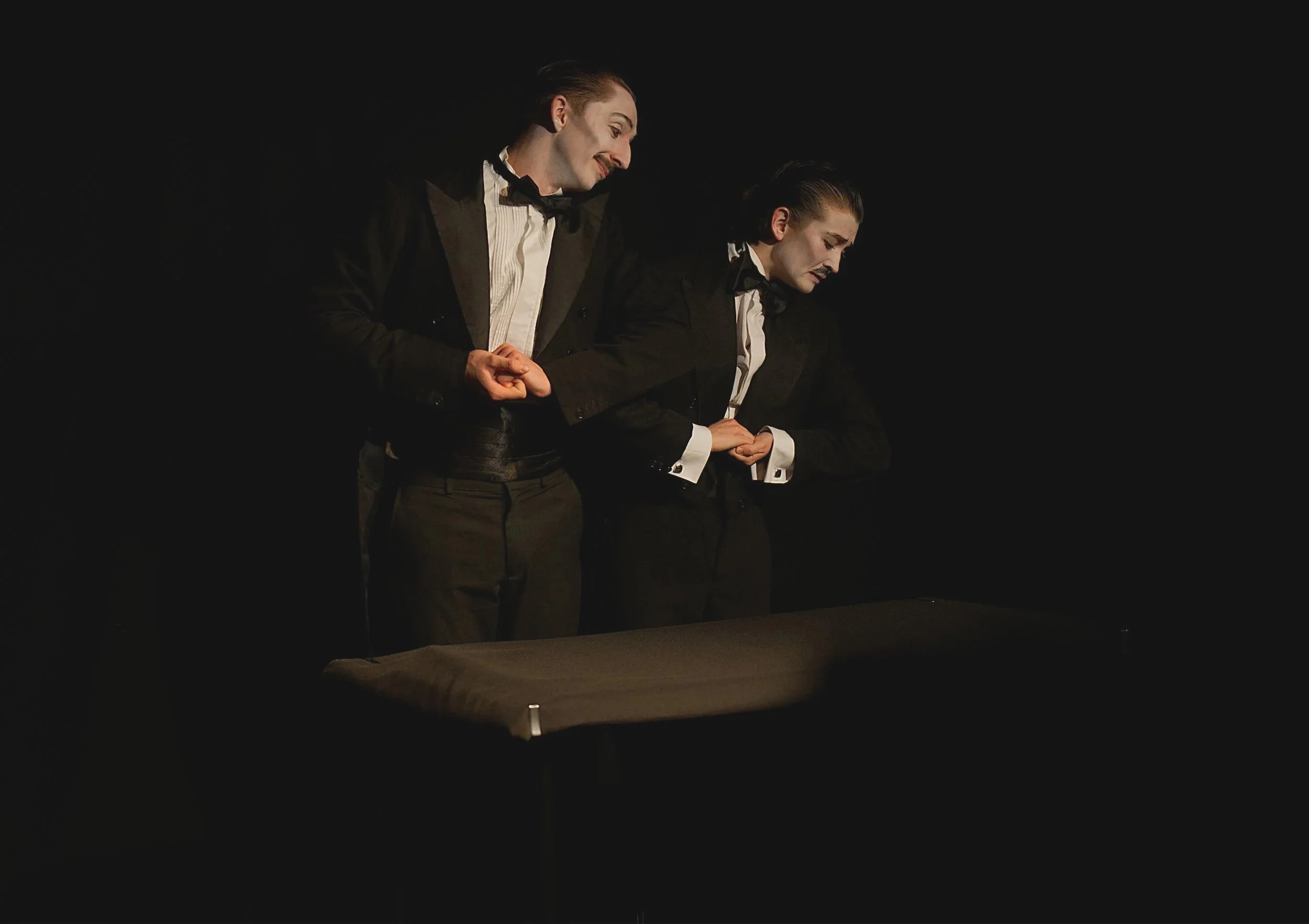 Two actors dressed as waiters standing behind a table, engaging in a theatrical performance, with a dark background.