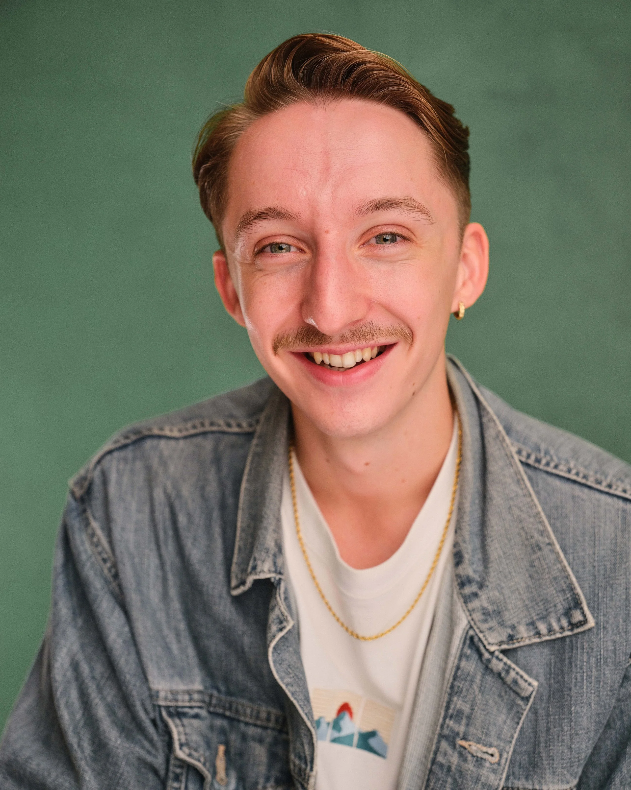 A young man with short brown hair, a mustache, and blue eyes smiling at the camera, wearing a dark green shirt against a dark background.