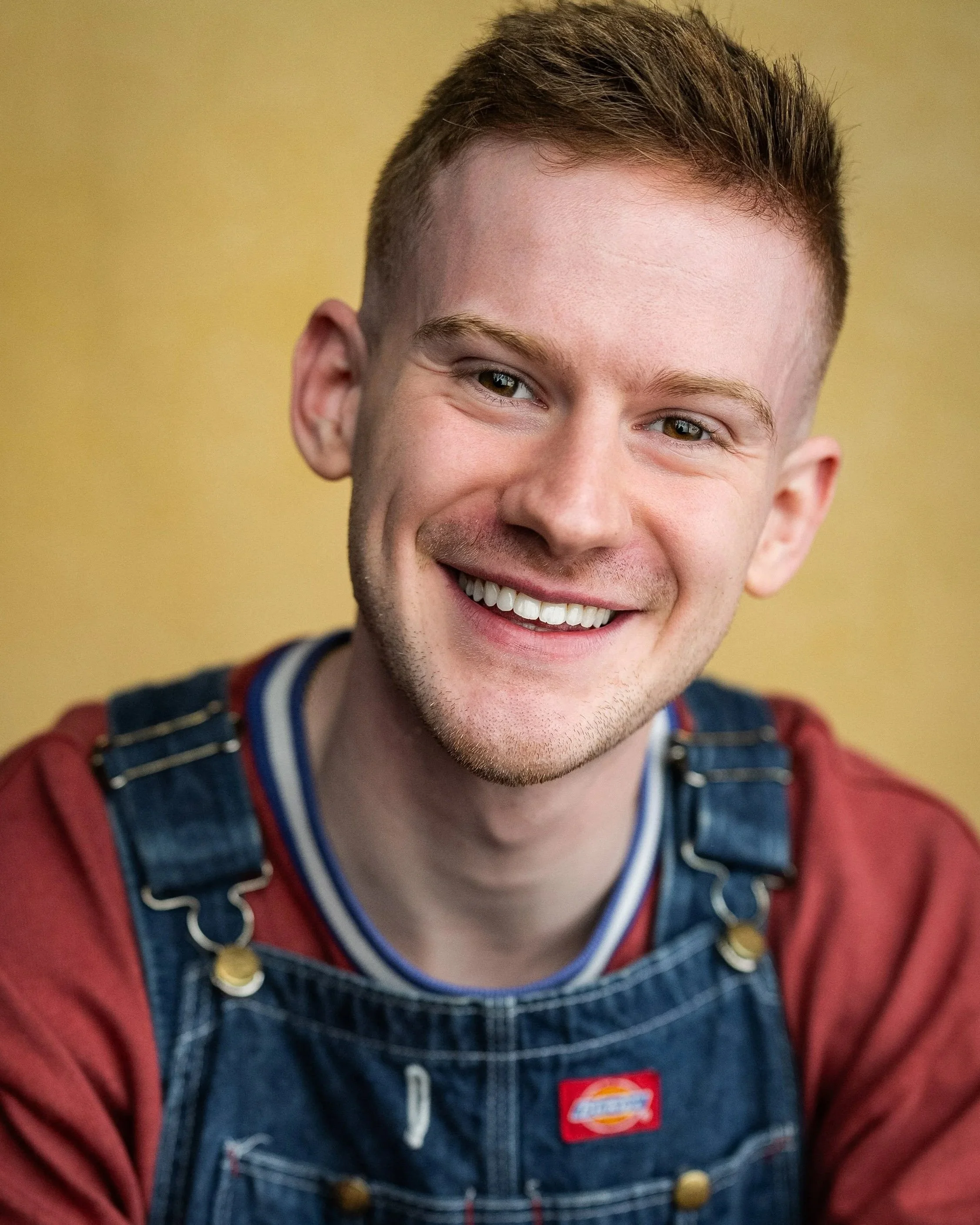 Headshot of a young man with short red hair, a beard, and wearing a blue denim shirt over a white T-shirt, smiling against a black background.