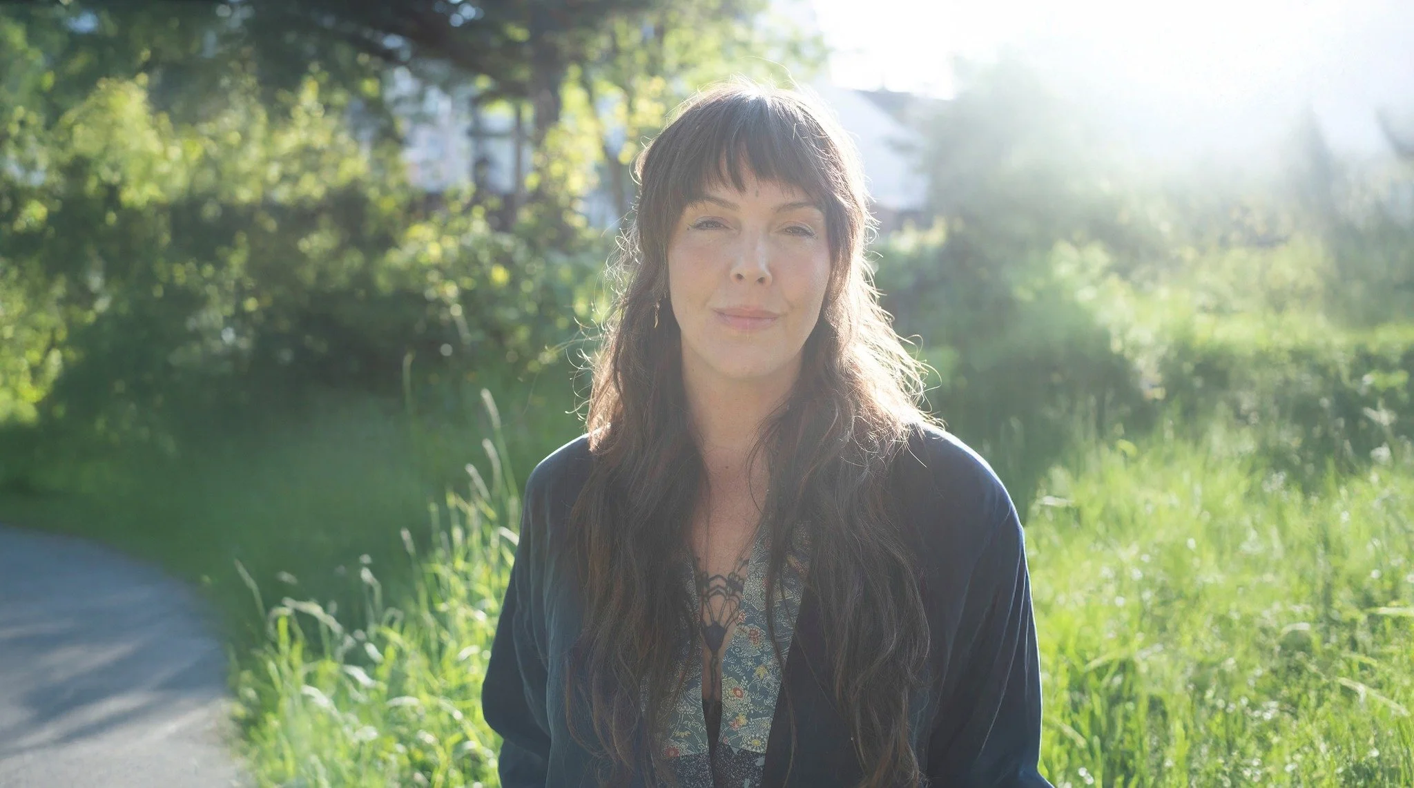 A woman with long wavy hair and bangs, standing outdoors in bright sunlight, near a grassy area with trees in the background.