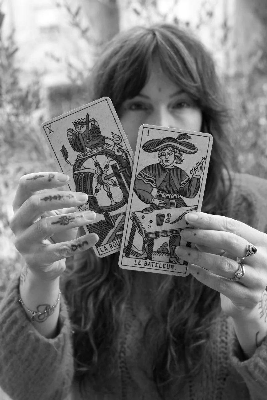 A woman with wavy hair holding two tarot cards, La Roue de Fortune and Le Batteleur, in front of her face outdoors, with trees in the background.