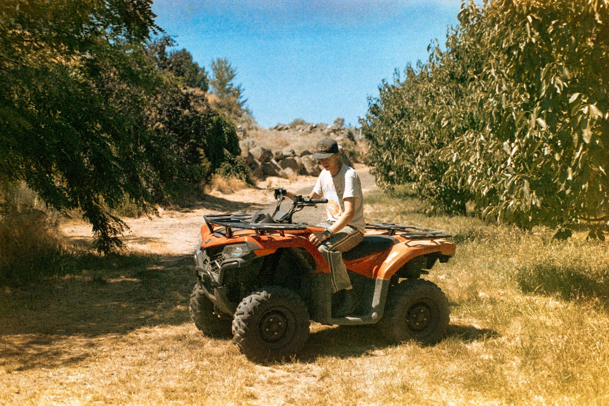 A person wearing a hat and white t-shirt, sitting on an orange all-terrain vehicle (ATV) on a dirt trail surrounded by greenery and trees, under a clear blue sky.