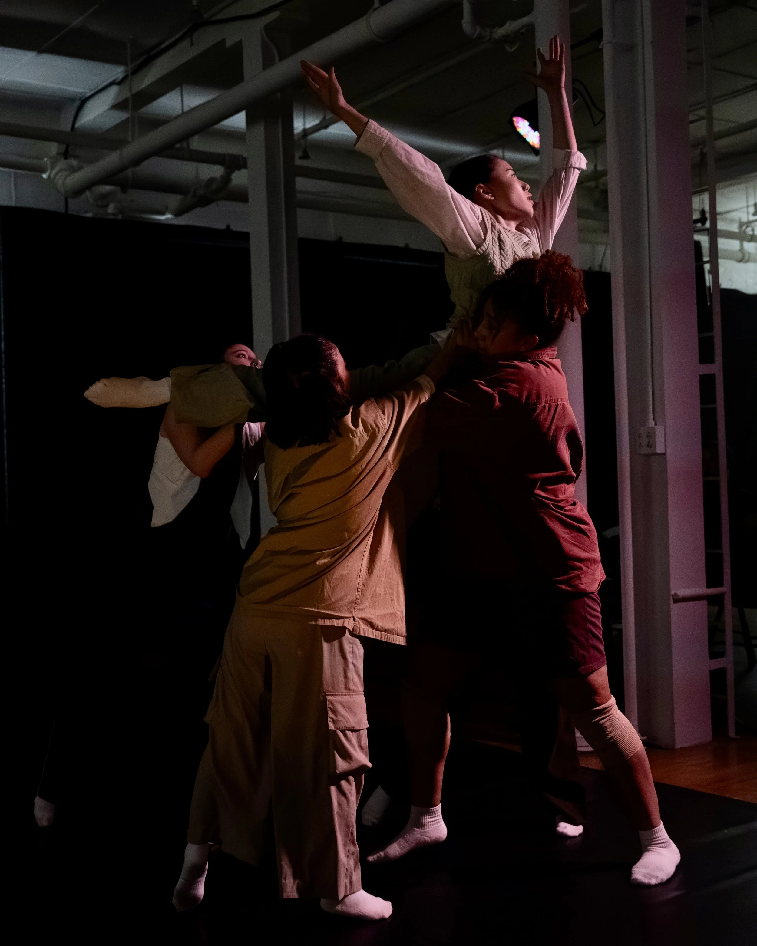 A group of four people lifting and supporting a woman reaching out to a spotlight in front of her, with one person holding her waist and others balancing her arms and shoulders, in a dark stage setting.