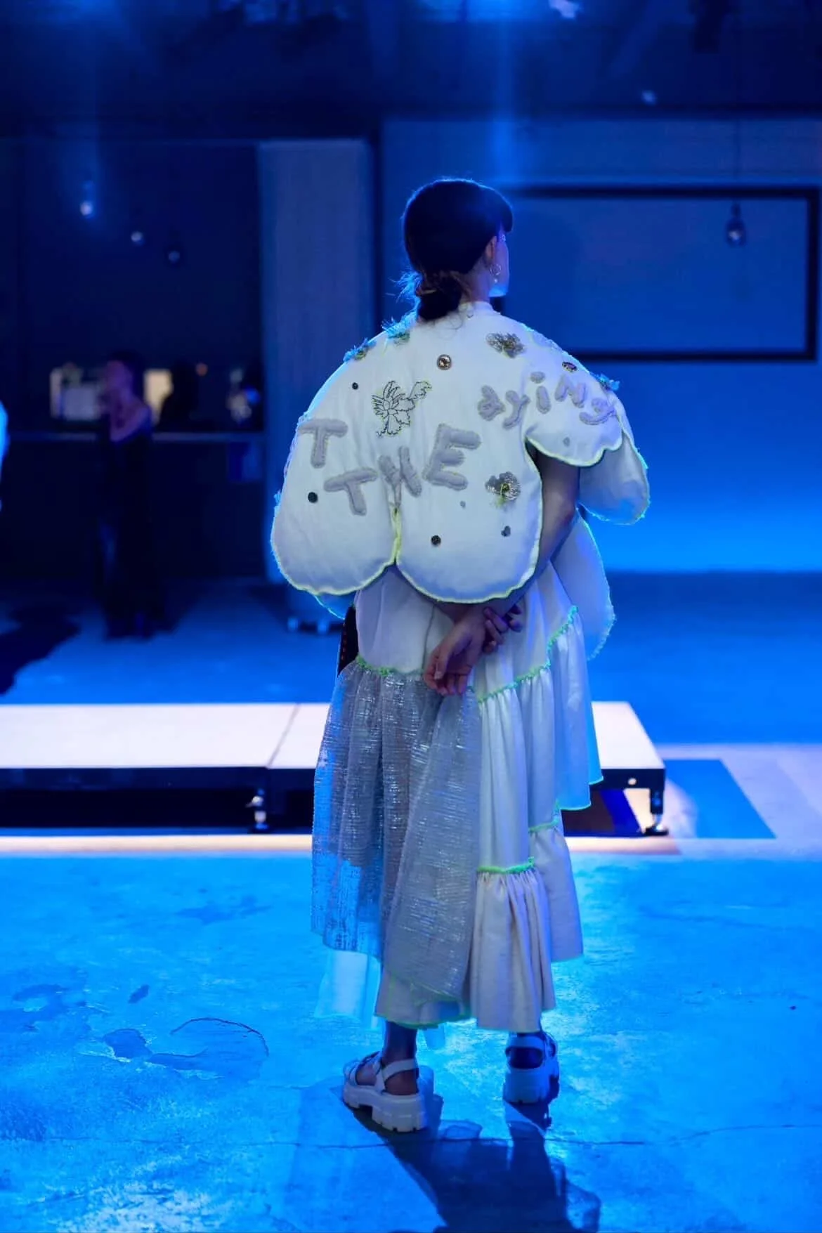 Person standing on a stage with a dark blue background, wearing a white puffy jacket with embroidered and beaded designs, and a long white skirt, seen from behind. The jacket has rounded edges and reads "when I left the dying".
