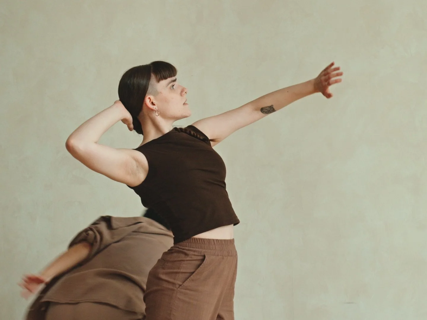 A dancer with braided hair, an undercut, and a tattoo on their forearm stretching their right arm forward against a plain light-colored wall. Their left hand is behind their head and they are wearing a brown, boat-necked top and brown pants.