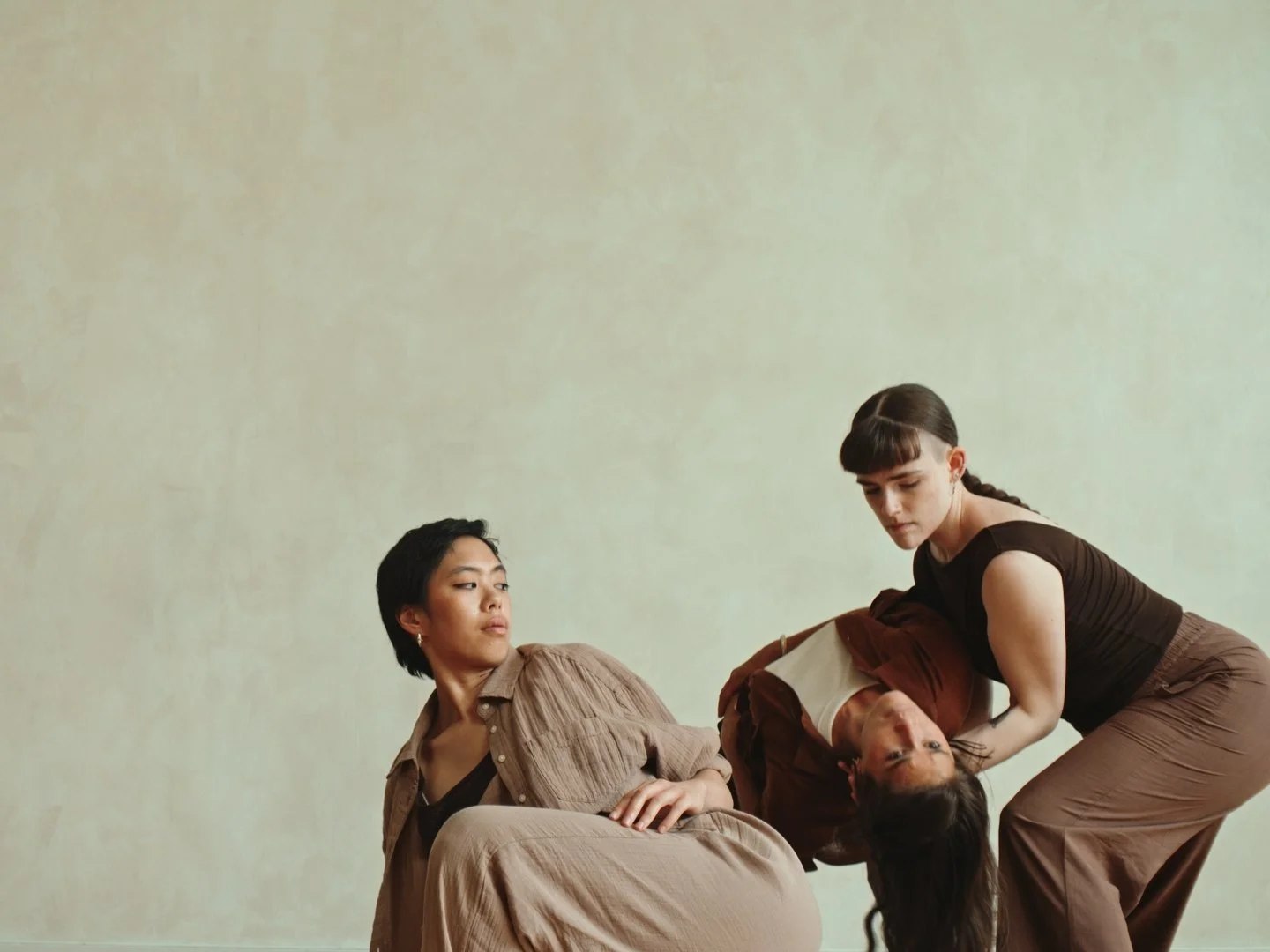 Three dancers in earthy-toned clothing, one seated and another leaning back into the third's arms, against a plain beige background.