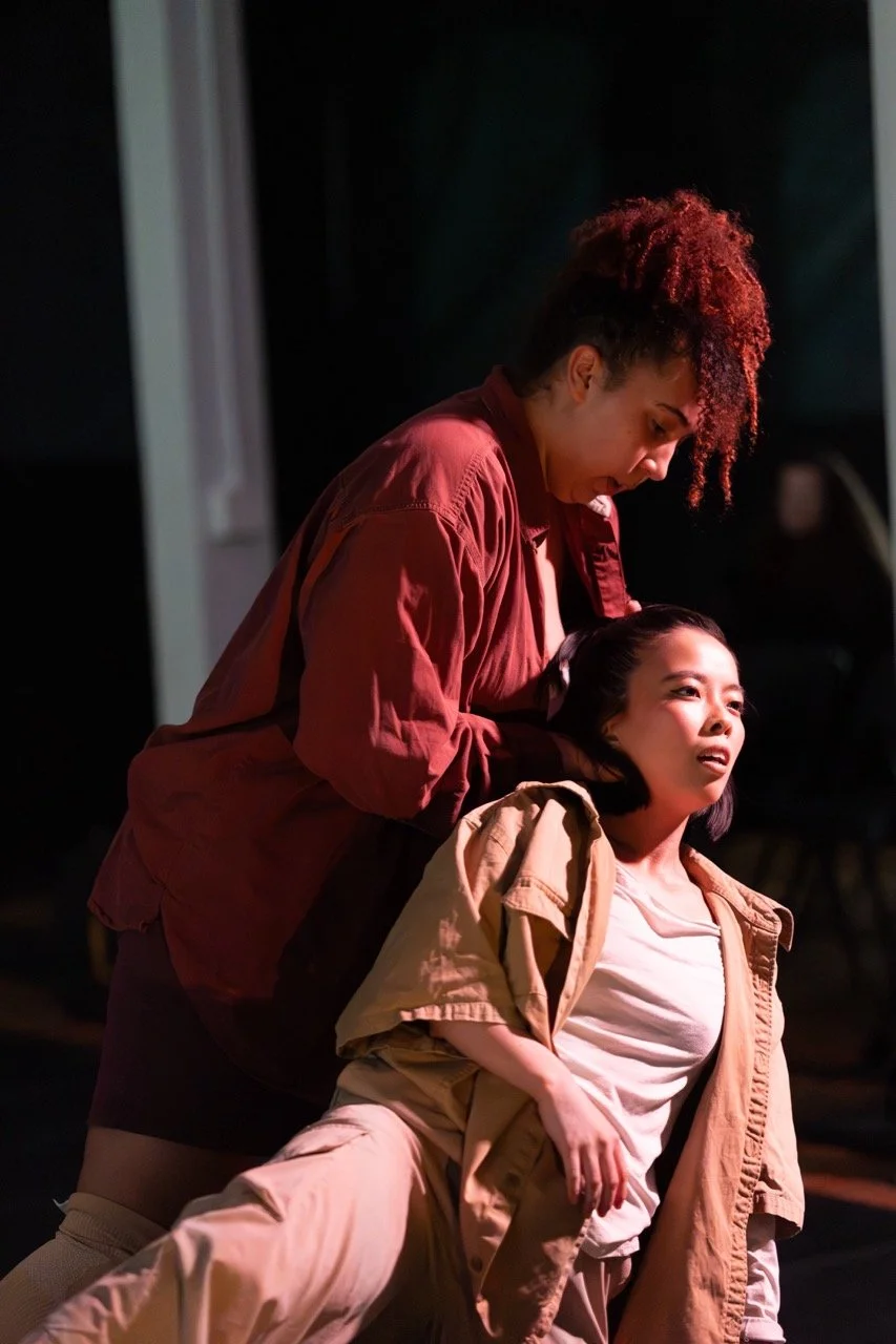 Two women on stage. One woman is standing, looking down with a concerned expression. The other's head is being supported from the side by the other's outstretched palms, looking forward blankly.