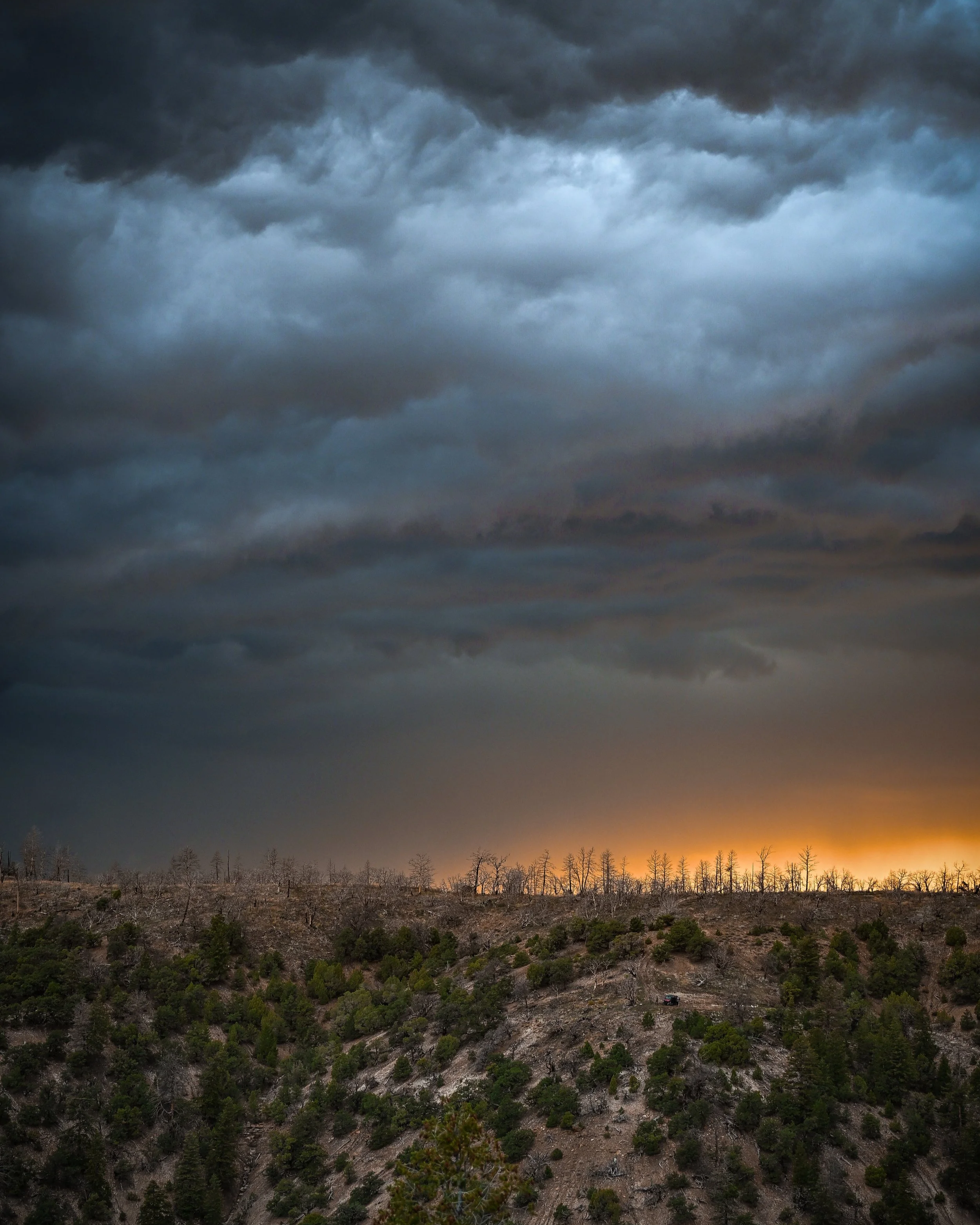 Dark storm clouds overhead as sunset lights the horizon, with a landscape of trees and a small vehicle in the distance.