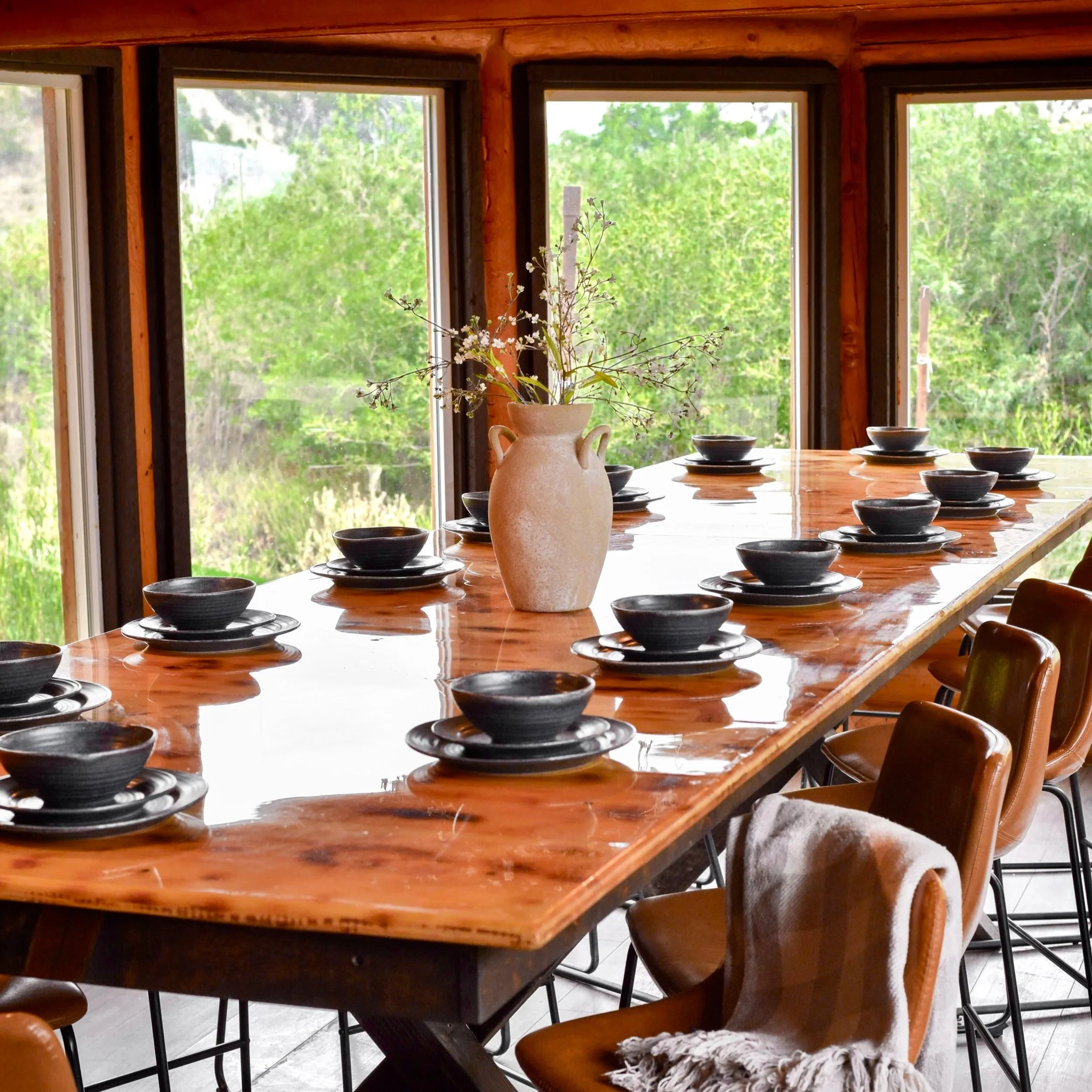 A wooden dining table with black bowls and a large beige vase with wildflowers, surrounded by wooden chairs, in front of large windows showing green trees outside.
