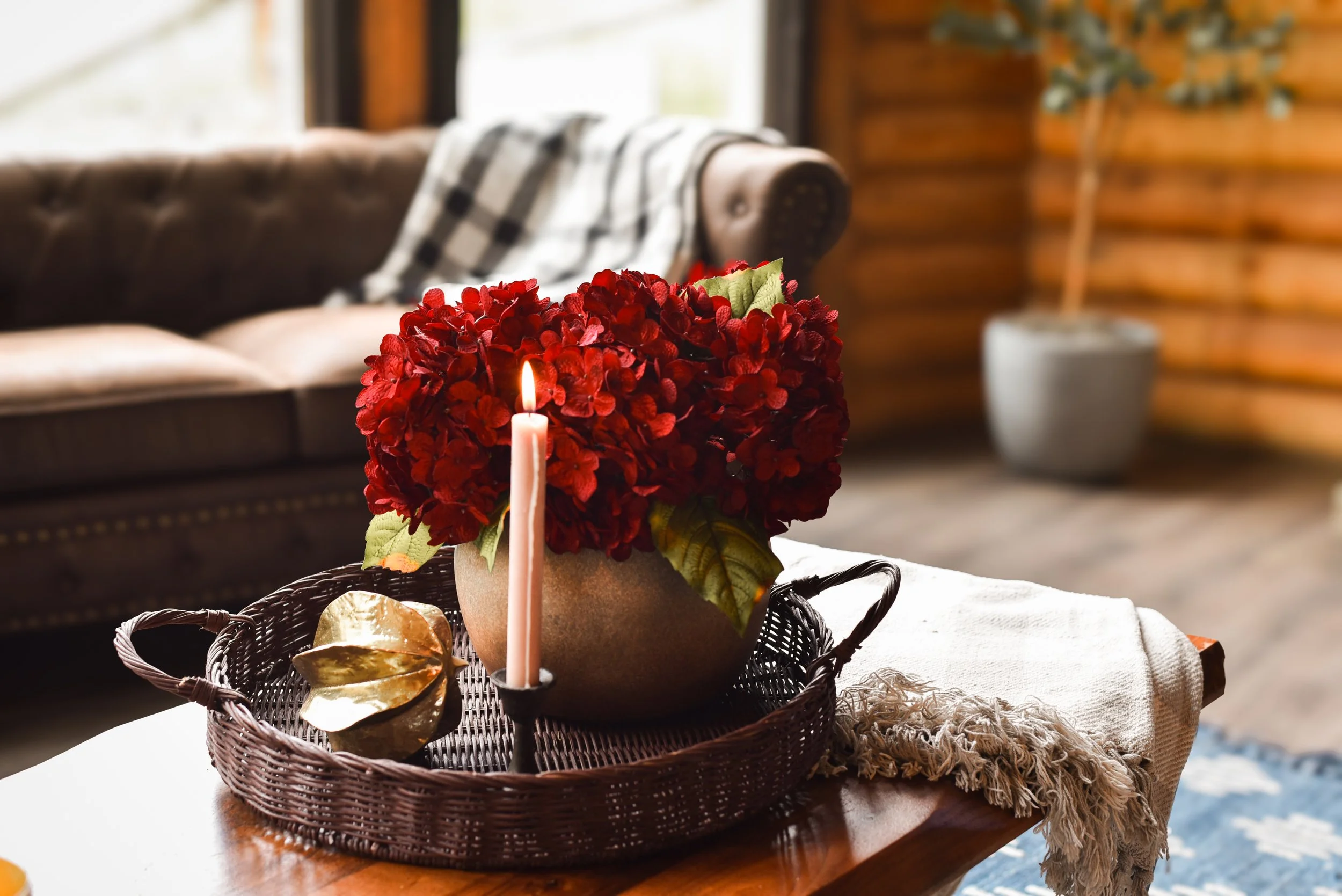 Decorative arrangement on a wicker tray with a pink candle, a gold decorative object, and a red floral arrangement, placed on a table in a cozy, rustic interior.
