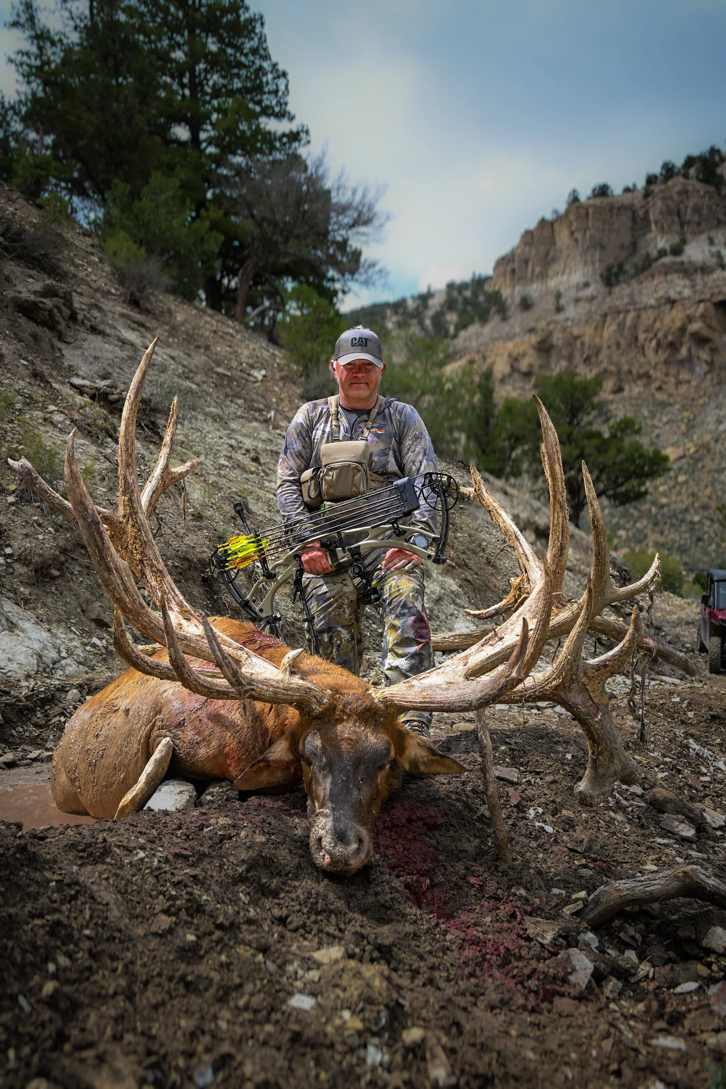 A man holding a bow and arrow, sitting behind a large elk with impressive antlers in a rugged outdoor landscape with trees and cliffs.
