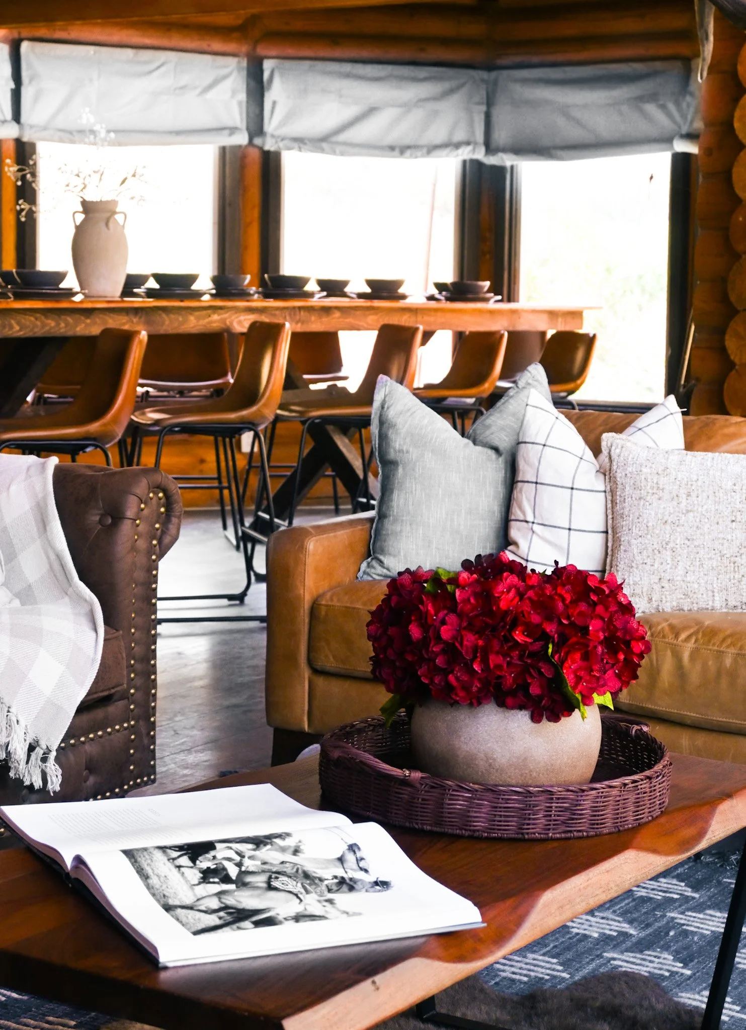 Cozy interior with a wooden dining table, chairs, decorative pillows, red flowers in a pot, and a book with black and white photographs on top.
