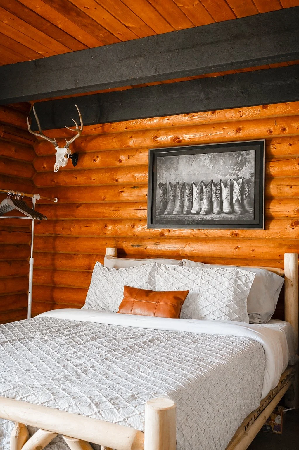 A rustic bedroom with wood-paneled walls and ceiling, a mounted deer skull with antlers, a framed black and white photo of cowboy boots, a bed with a white quilt and pillows, and a small clothes rack with hangers.