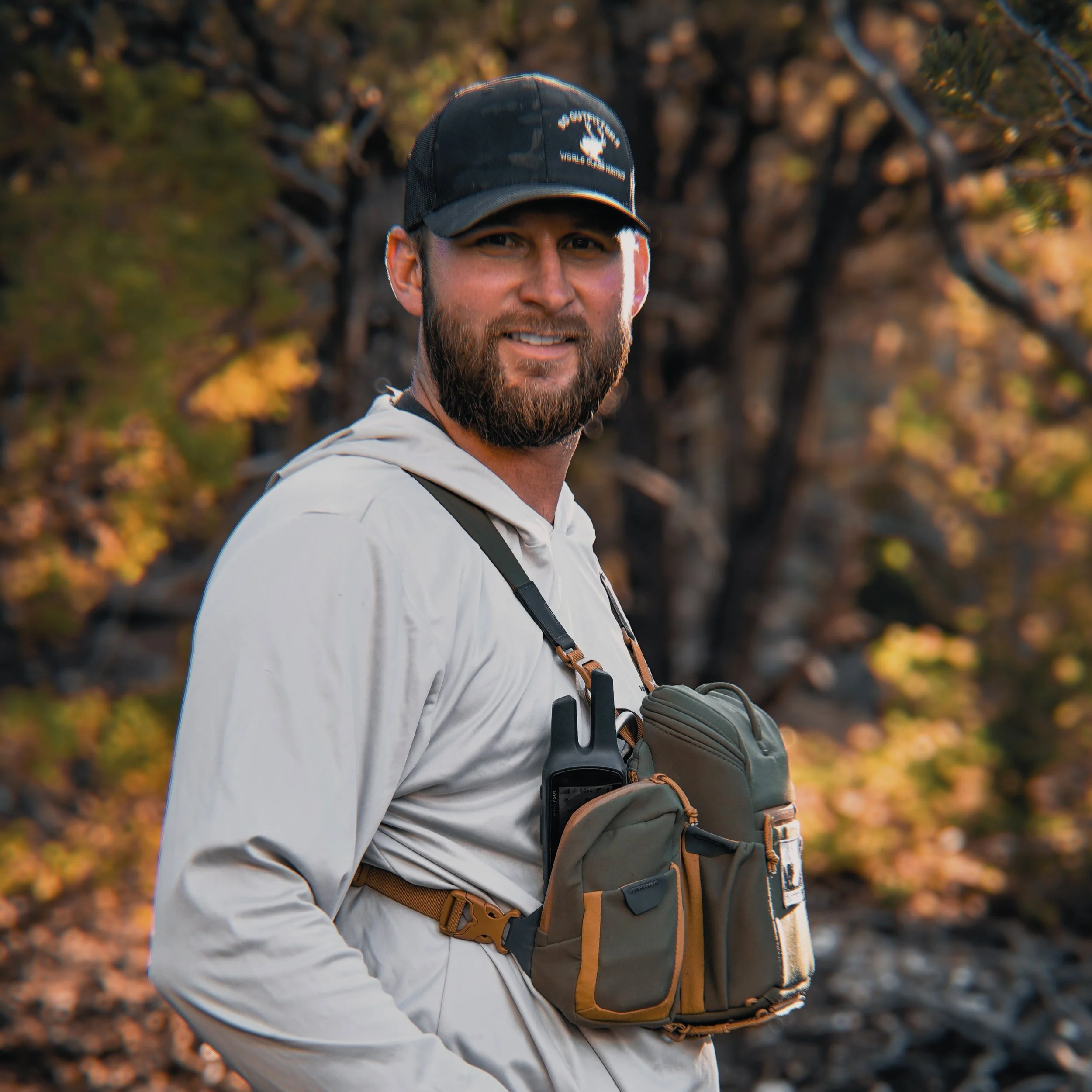A man with a beard wearing a black cap that says 'GUTREND' and a white jacket, standing outdoors in front of blurred trees with fall foliage.