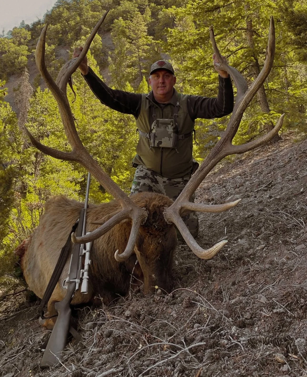 A man in outdoor clothing and a cap holding up antlers of a large elk with a rifle lying on the ground beside it in a wooded area.