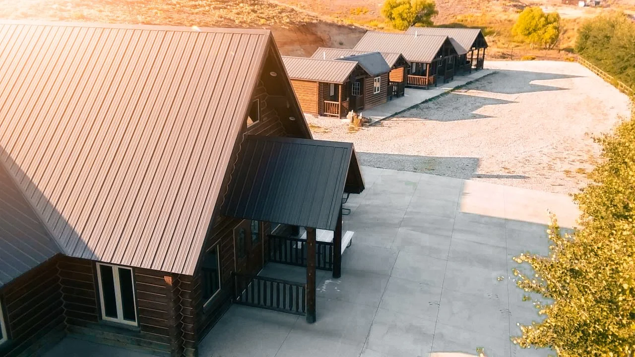 Aerial view of multiple small wooden cabins on a gravel lot with a large concrete area and trees with autumn foliage in the background.