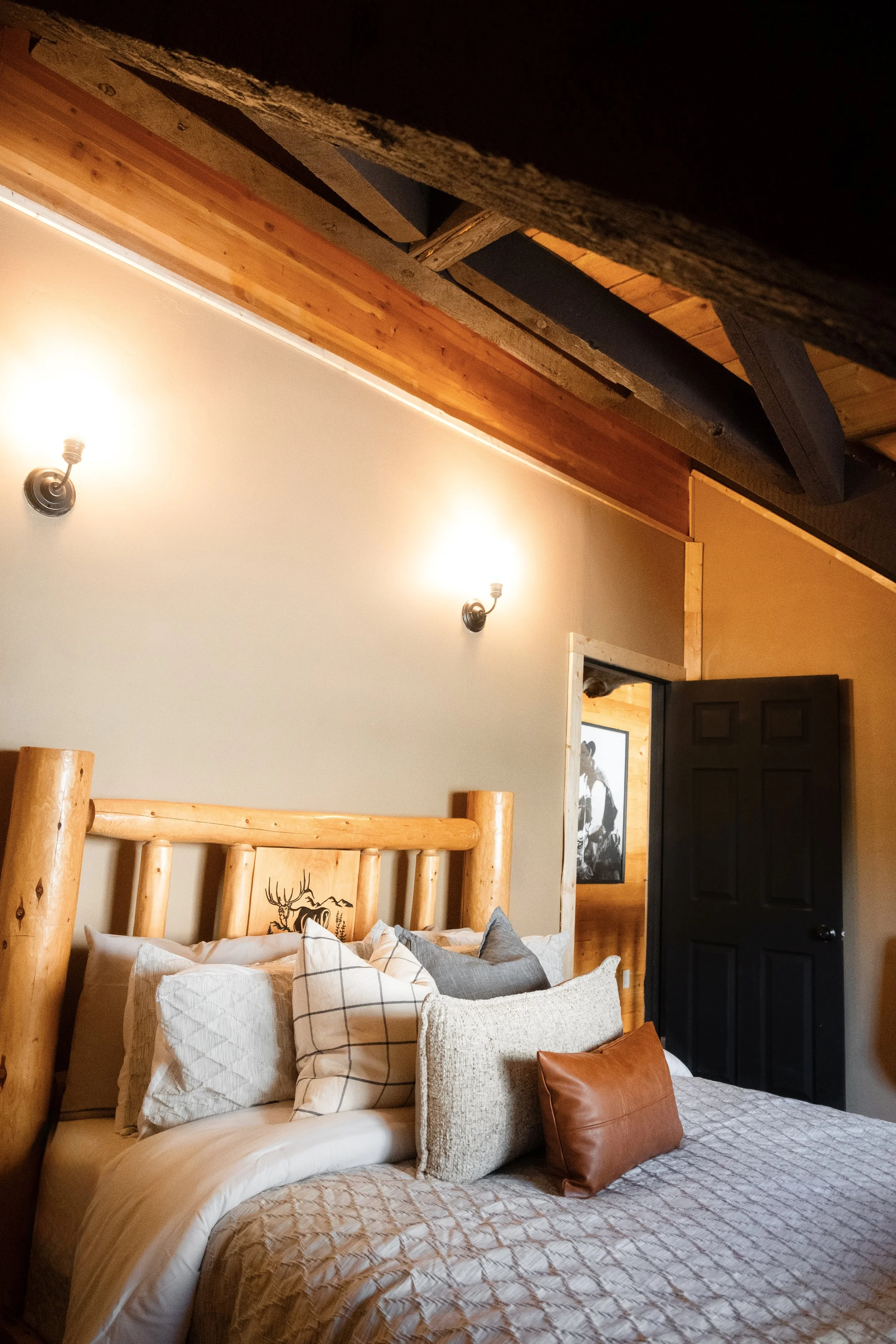 A cozy bedroom with a log cabin style, featuring a wooden log bed frame, multiple pillows including a plaid one and a brown leather one, and a quilted beige bedspread. There are two wall-mounted light fixtures, a black door, and a framed black and wh