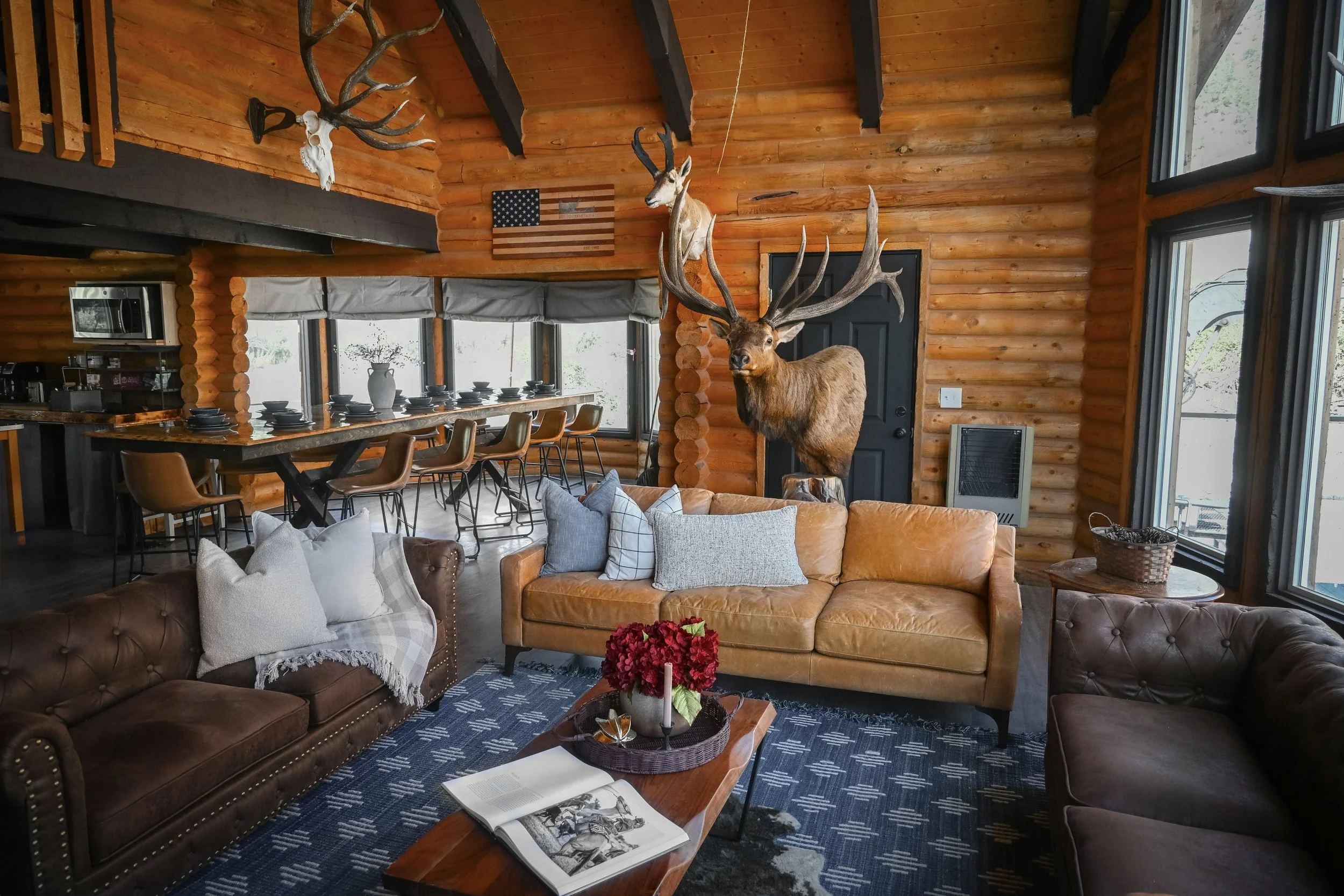 Interior of a log cabin living room with mounted deer and antelope heads on the wood-paneled walls, large windows, a brown leather sofa with pillows, a wooden coffee table with a magazine and a vase of flowers, and a dining area in the background wit