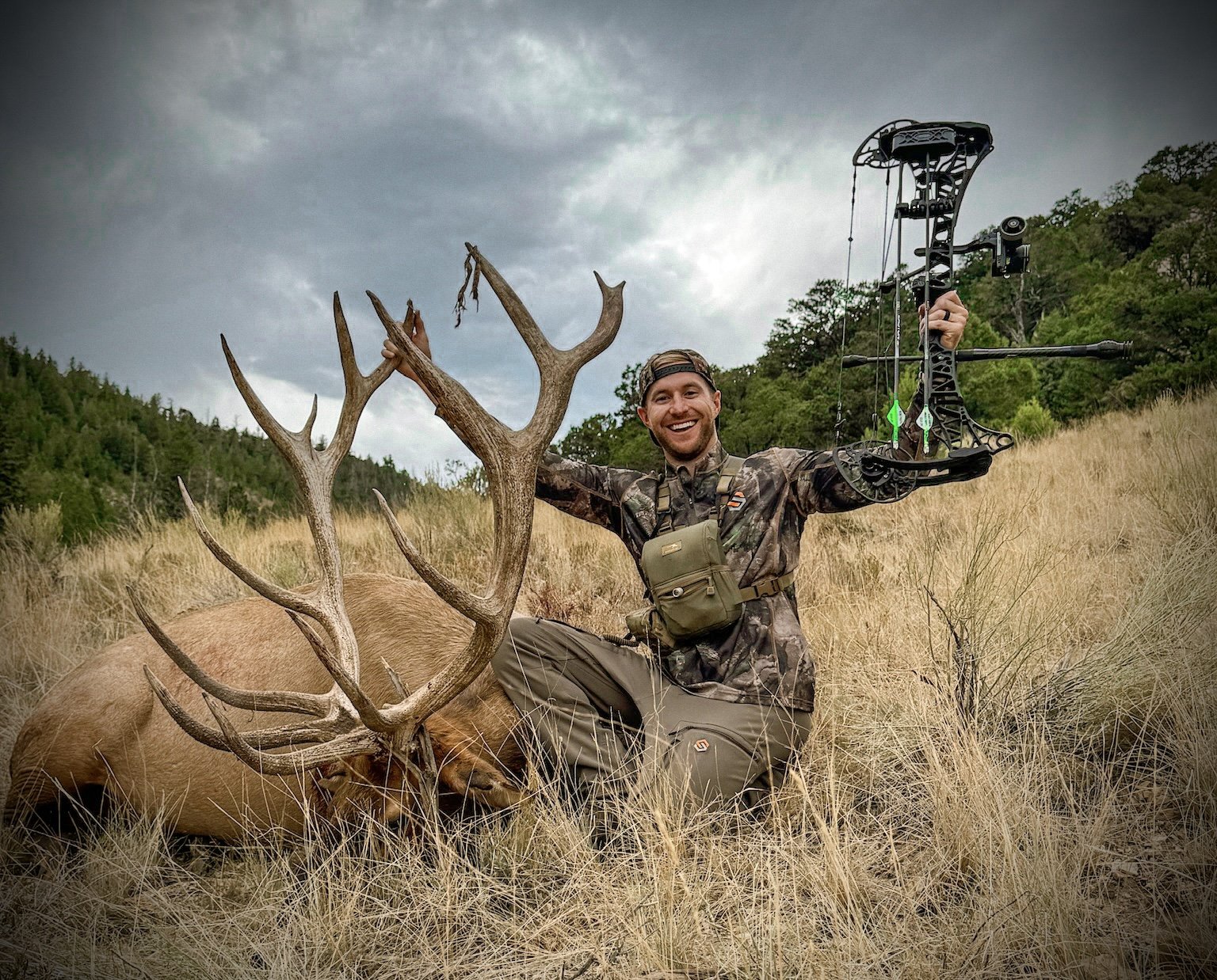 A man kneeling in a grassy field holding a bow with a hunted elk with large antlers beside him, overcast sky in the background.