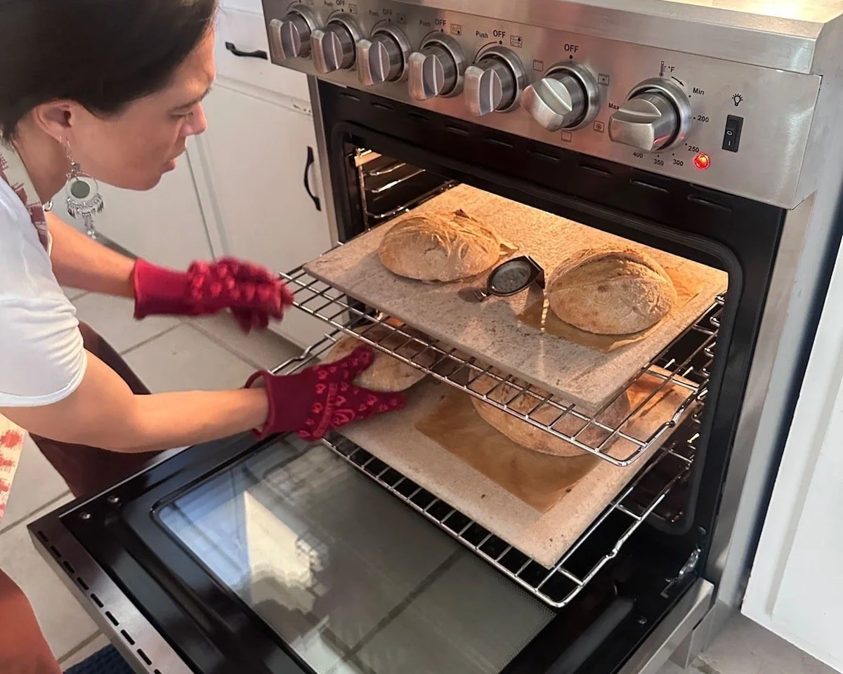 Woman using an oven to bake bread, with two loaves on baking sheets and a thermometer inside the oven.