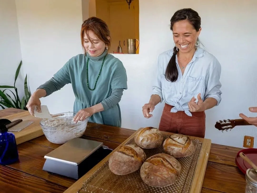 Two women standing at a wooden table with freshly baked bread and a bowl, one woman is mixing something in the bowl, the other is smiling and standing over freshly baked bread rounds, in a bright kitchen with a plant and a wall in the background.