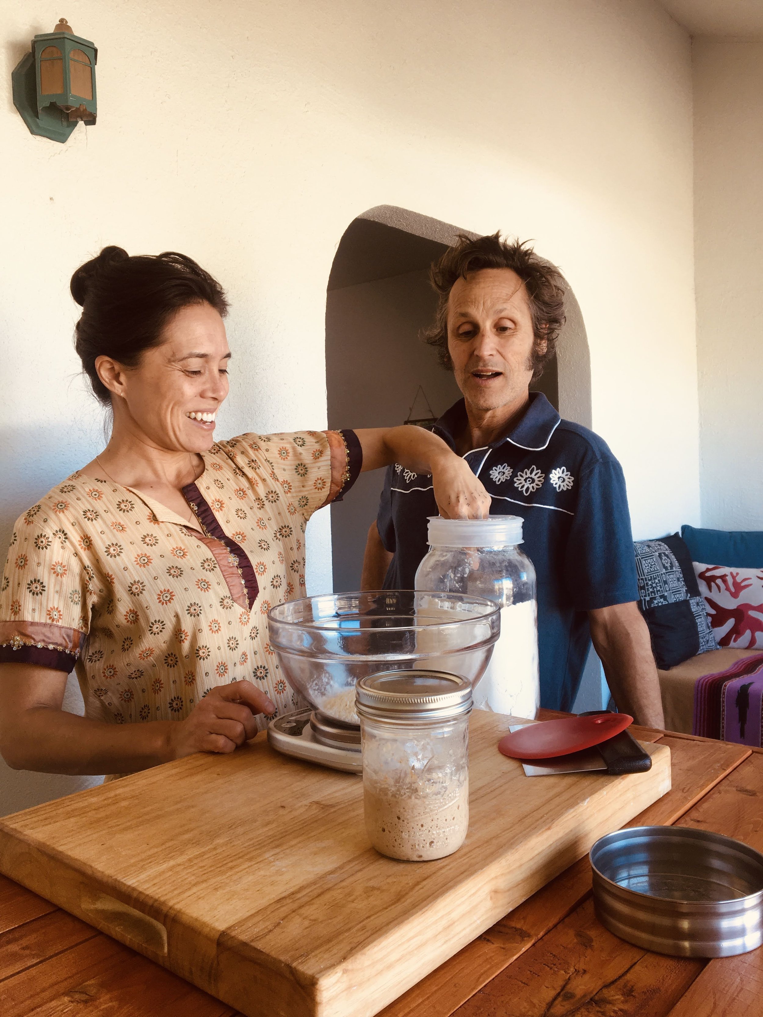 A woman smiles while measuring out flour on her kitchen table as her husband happily observes