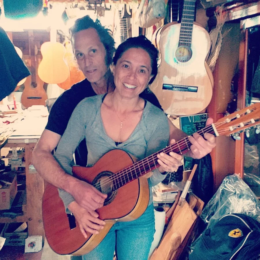 A young couple in a luthier shop with multiple guitars hanging on the wall; one woman is holding a guitar and smiling, her husband stands behind her, smiling.