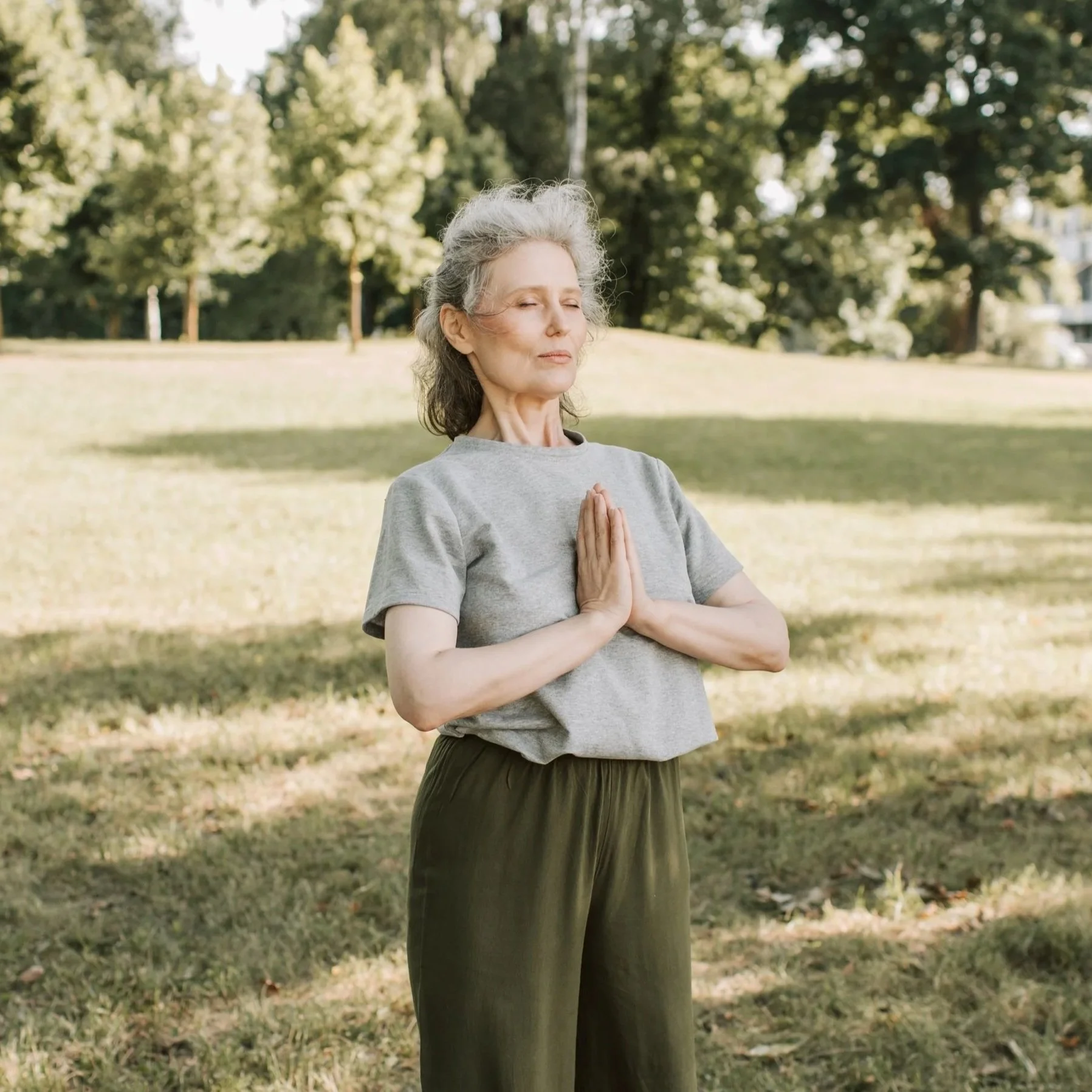 An elderly woman practicing yoga outdoors in a park, standing with her hands pressed together in a prayer position near her chest, eyes closed, wearing a gray t-shirt and dark pants, with trees and grass in the background.