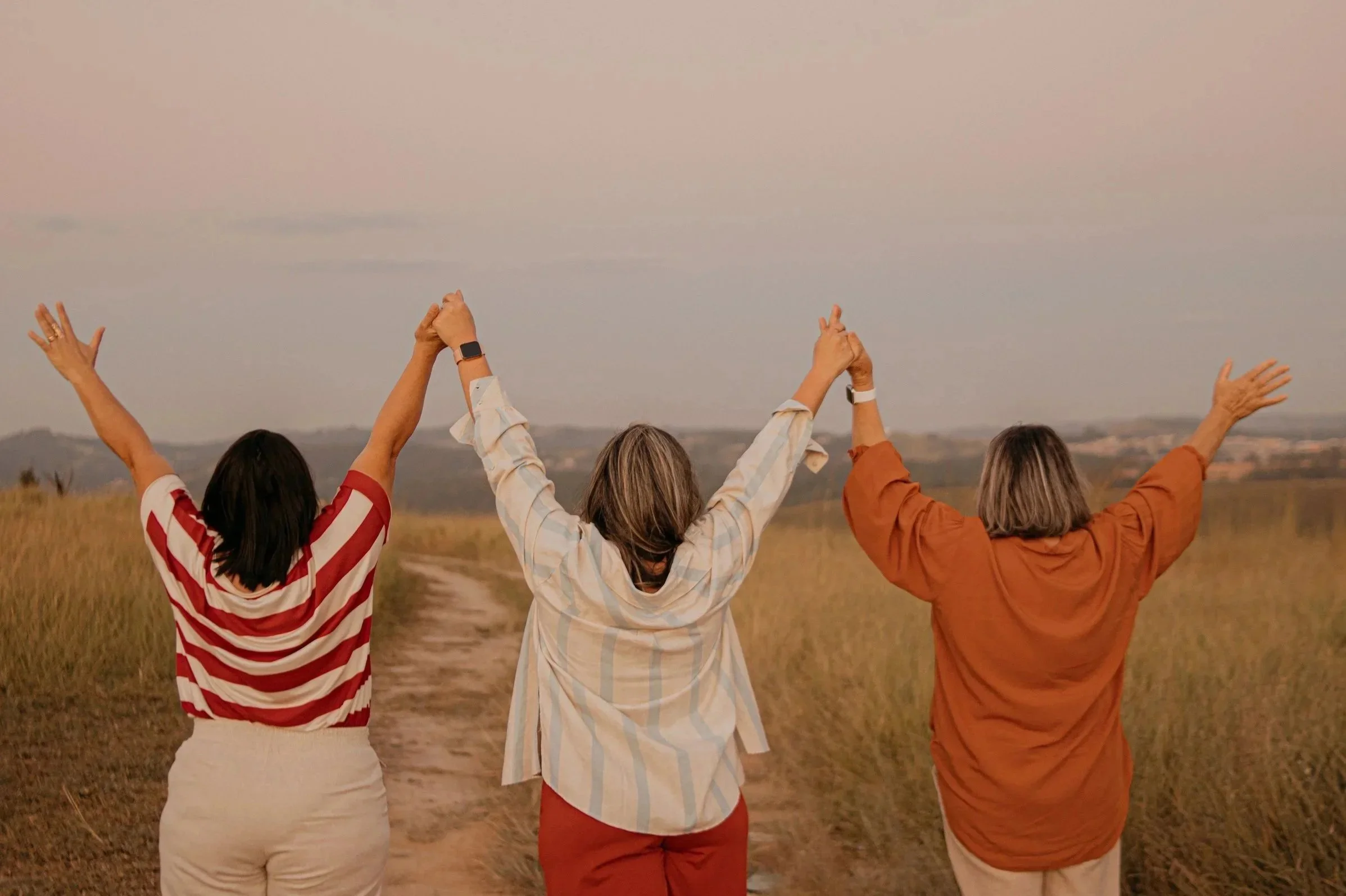 Three women standing outdoors in a field with their backs to the camera, raising their hands towards the sky during sunset.