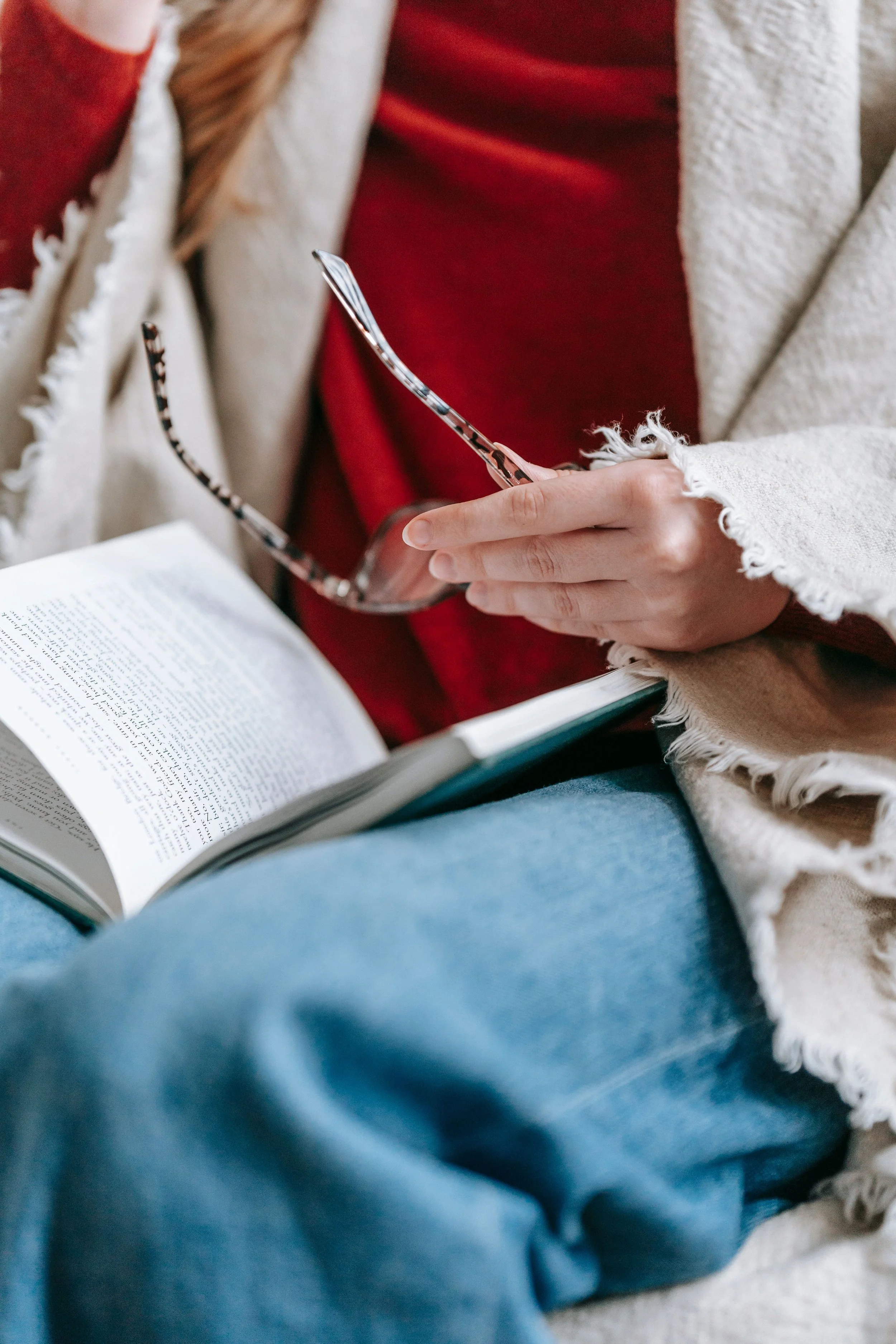 Person wearing blue jeans and a beige cardigan reading a book and holding glasses