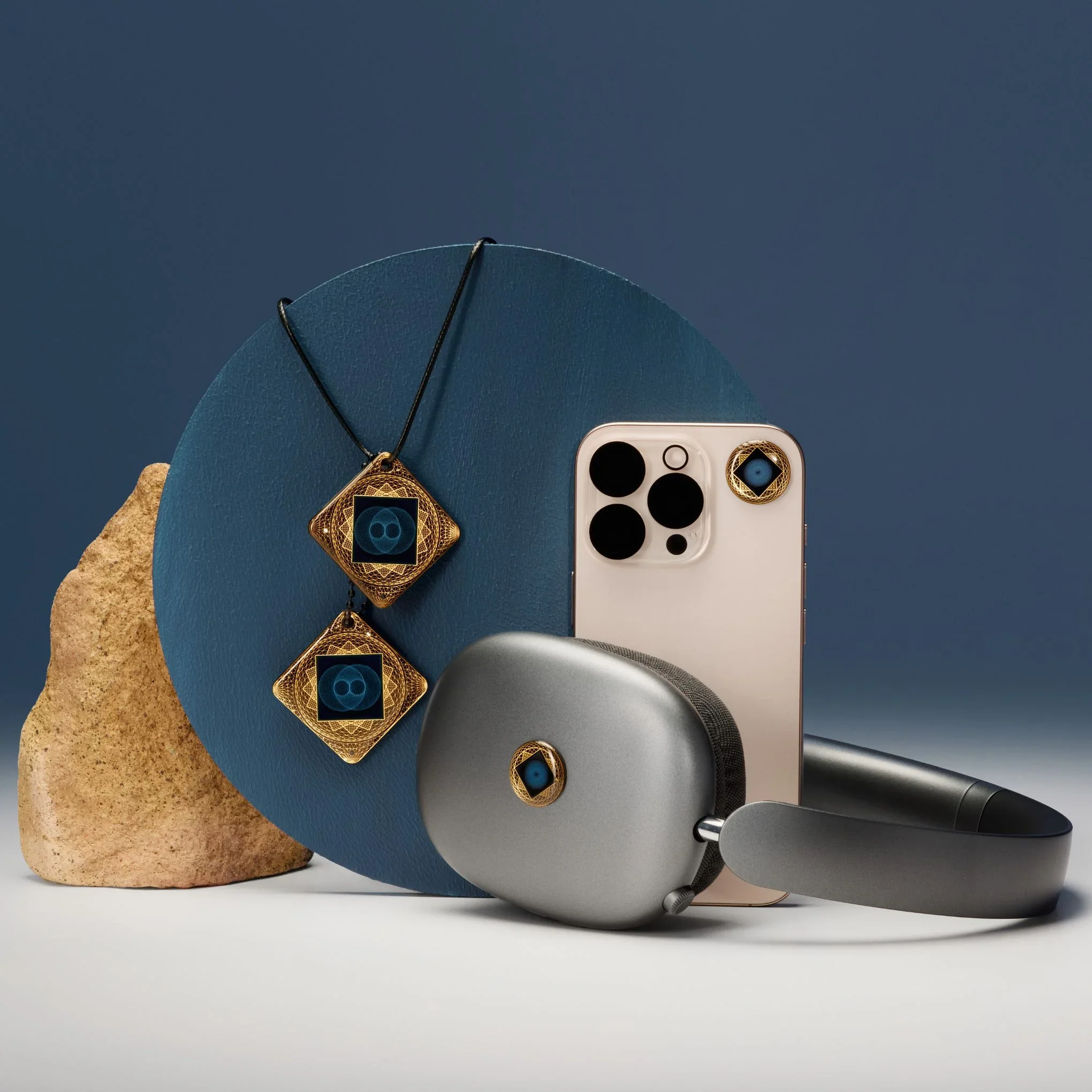 A smartphone with a white back and black camera lenses, a necklace with geometric pendants, a small speaker, a gray bag, and a magazine with circular designs, all placed on a blue circular surface against a dark blue background.