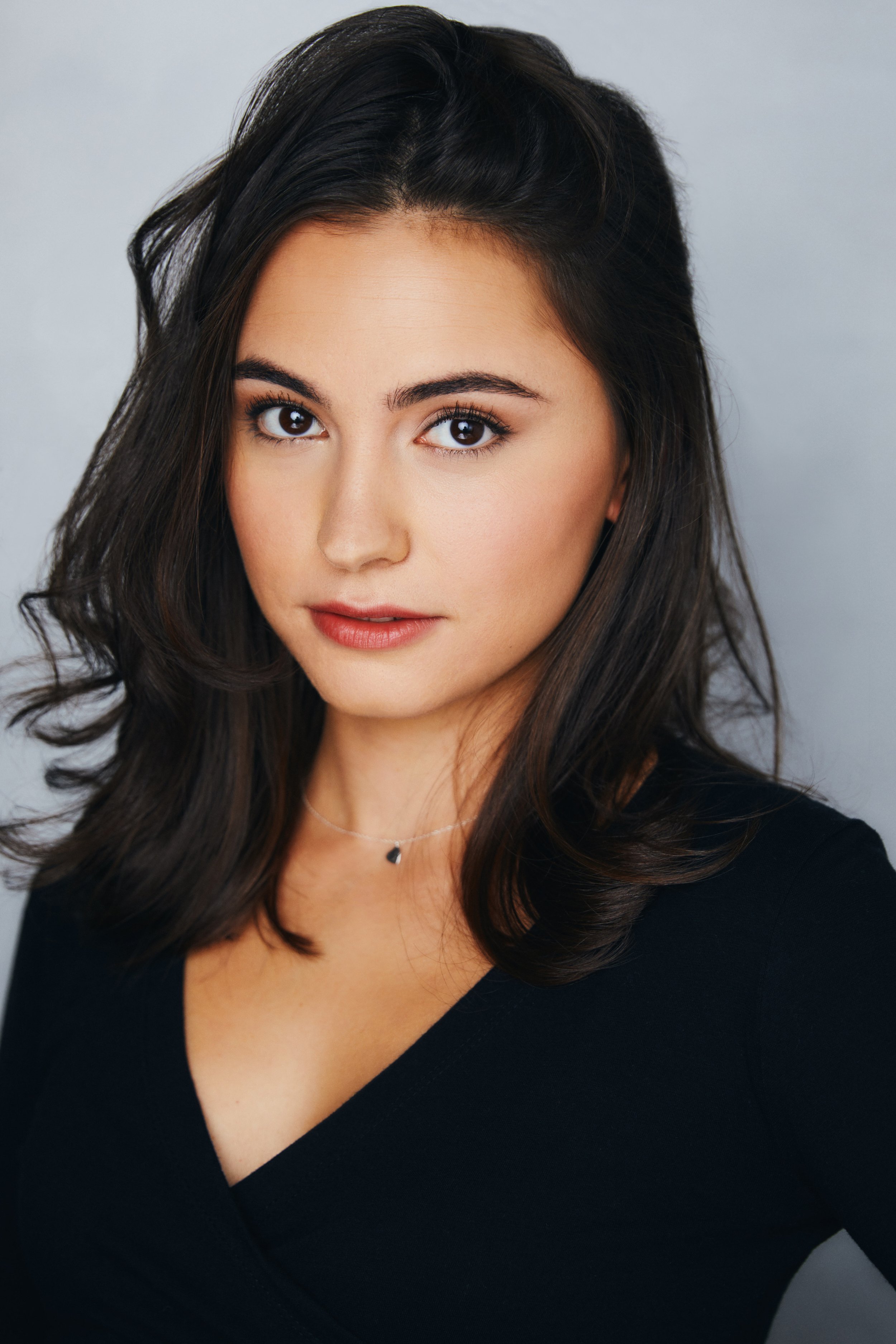 Portrait of a woman with dark brown hair, looking directly at the camera, wearing a black top and a delicate necklace, against a plain gray background.