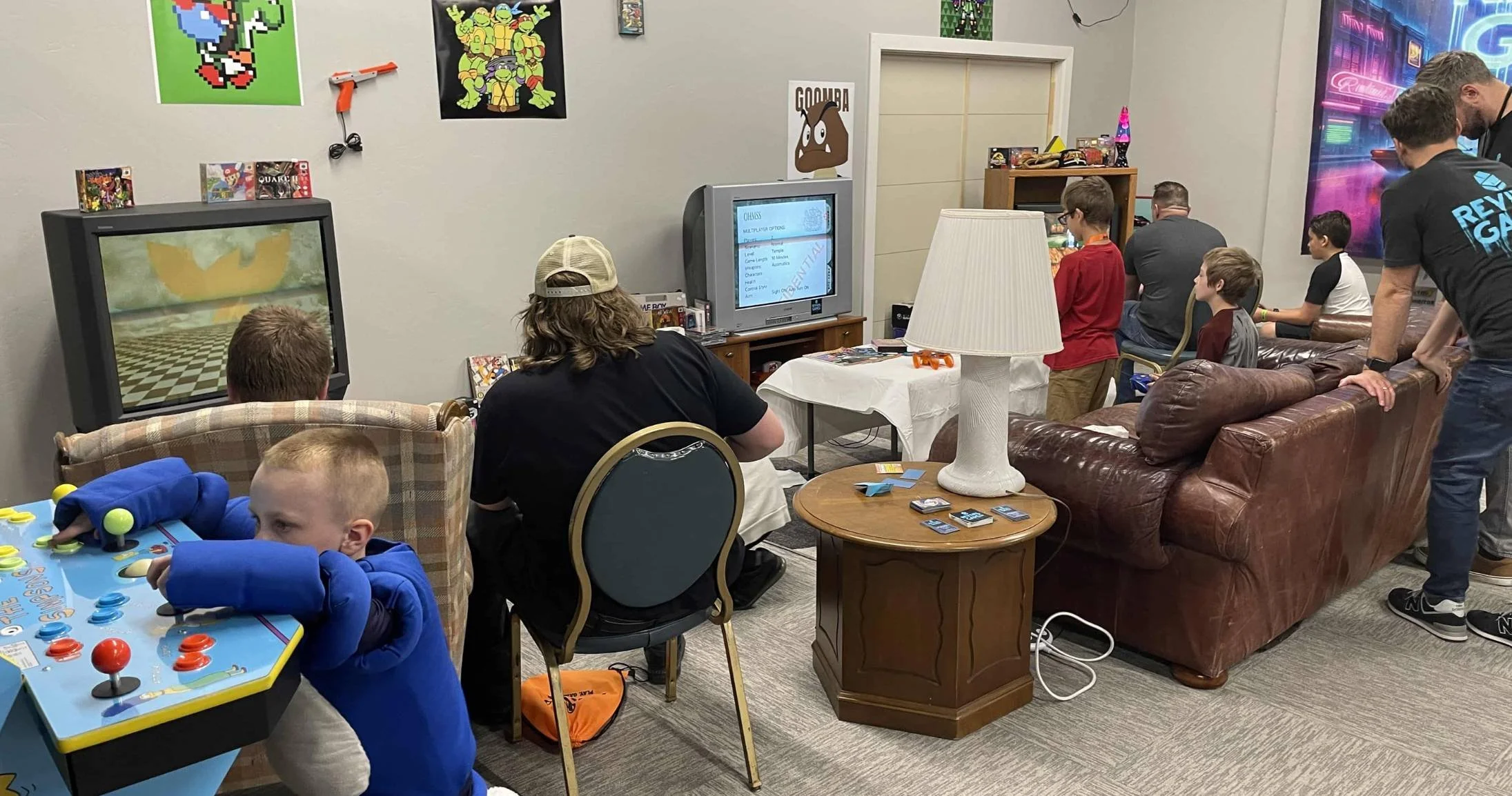 Picture from the game room at Retro X. Gamers are sitting on a brown couch in front of a console. Others are in chairs. Some people are looking on. The room has cream-colored carpet and walls.